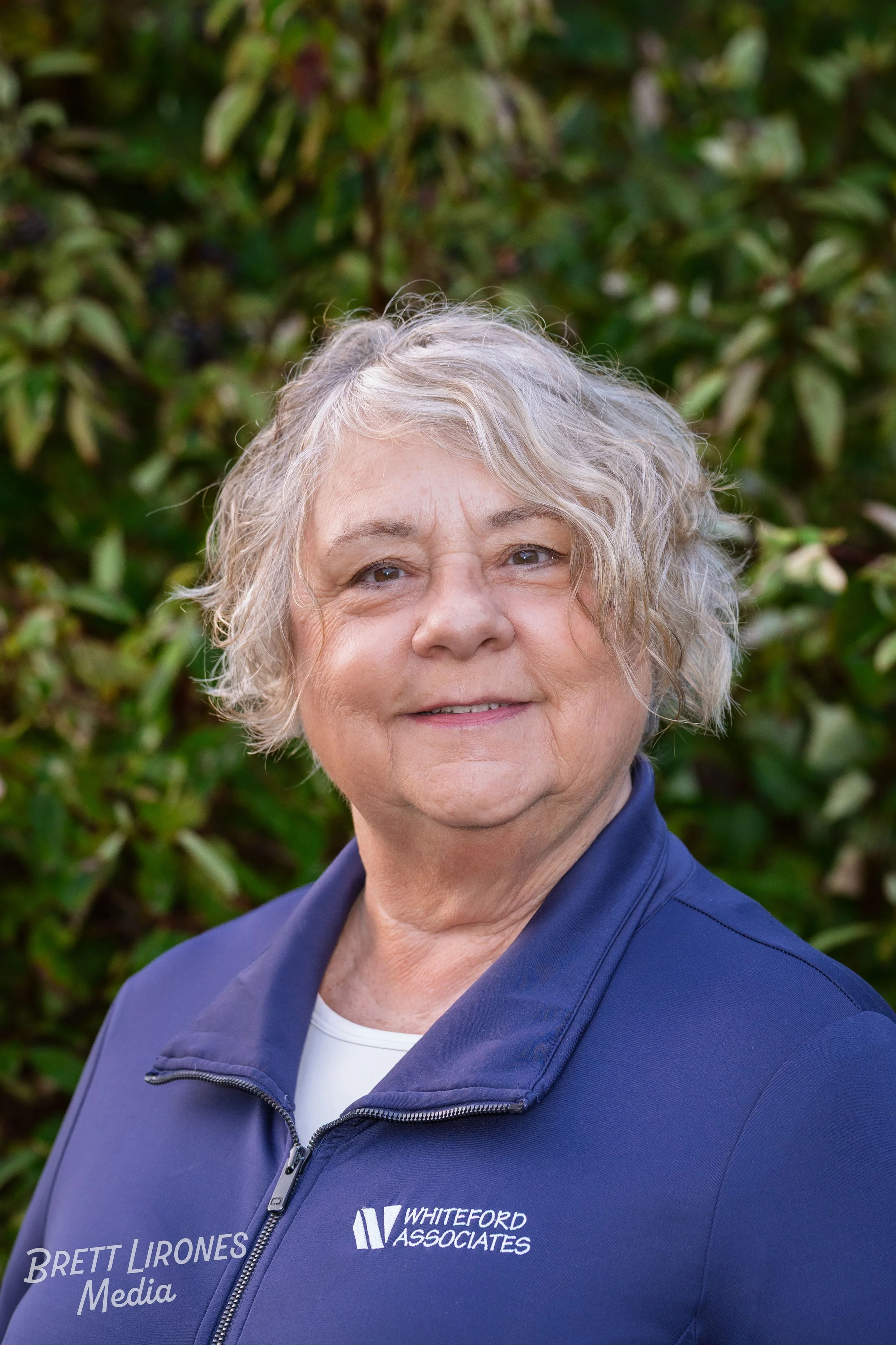 A smiling woman with short, wavy gray hair, wearing a blue jacket with the logo 'Whiteford Associates' and the text 'Brett Lirones Media,' standing outdoors in front of green foliage.