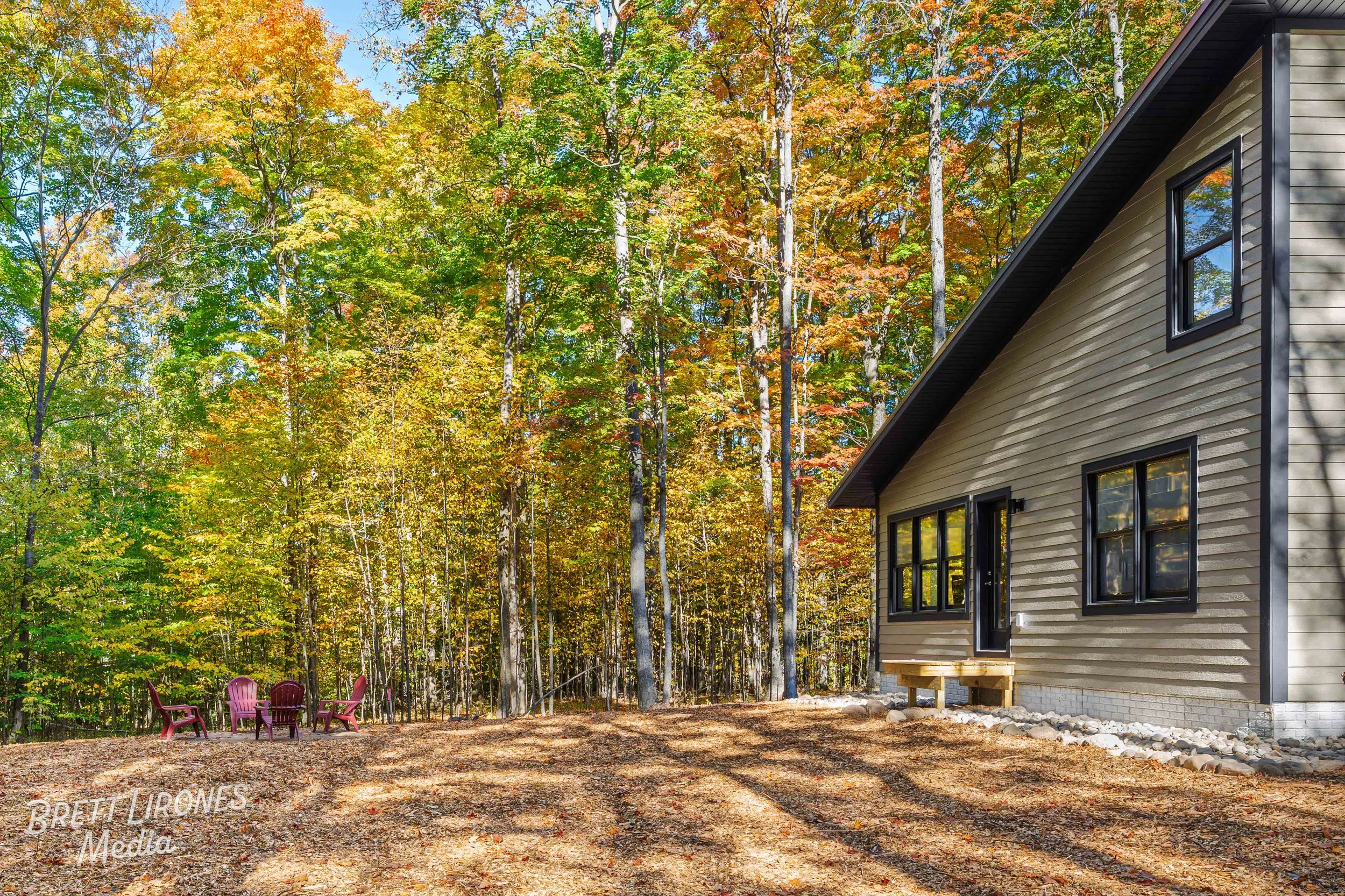 A house with beige siding and black-framed windows in a wooded area during fall with colorful trees and a small seating area with four pink chairs.