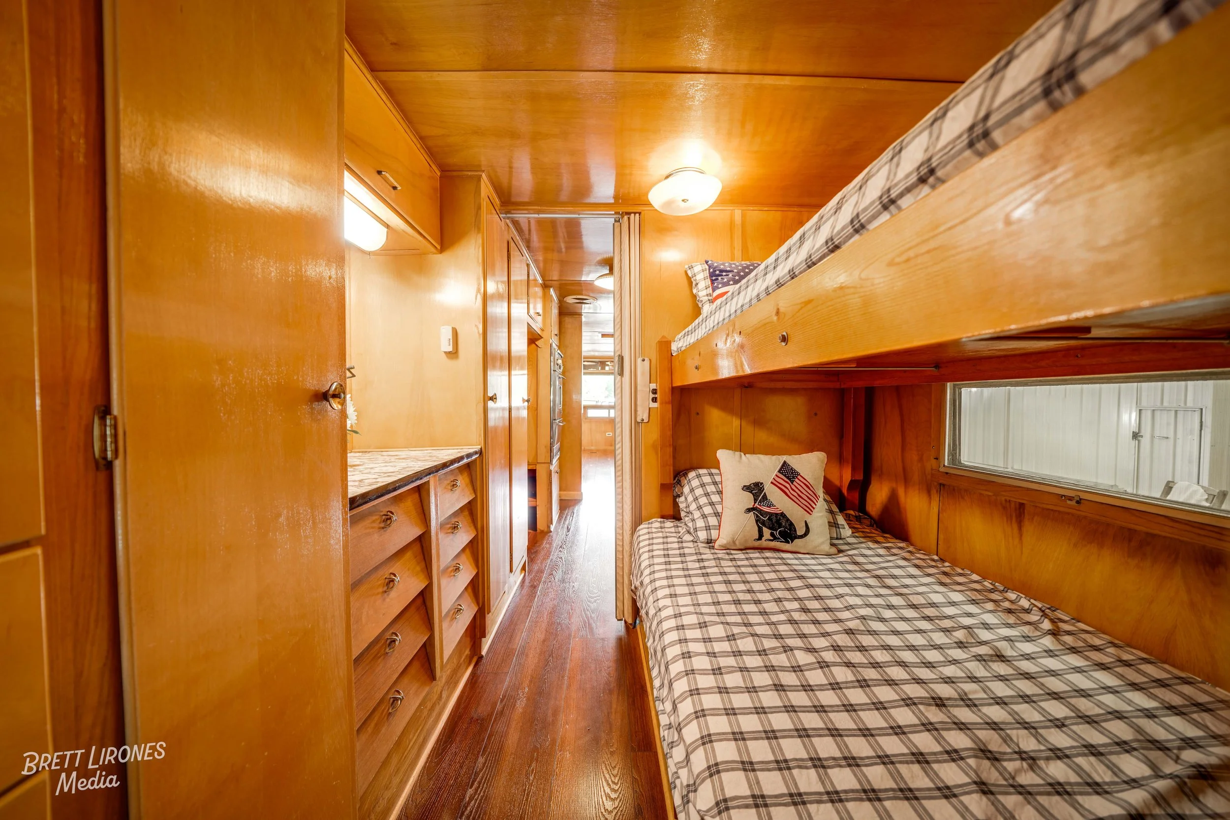 Interior of a wooden bunk bed room with two bunk beds, pillows, and a window, with a hallway leading to a kitchen area.