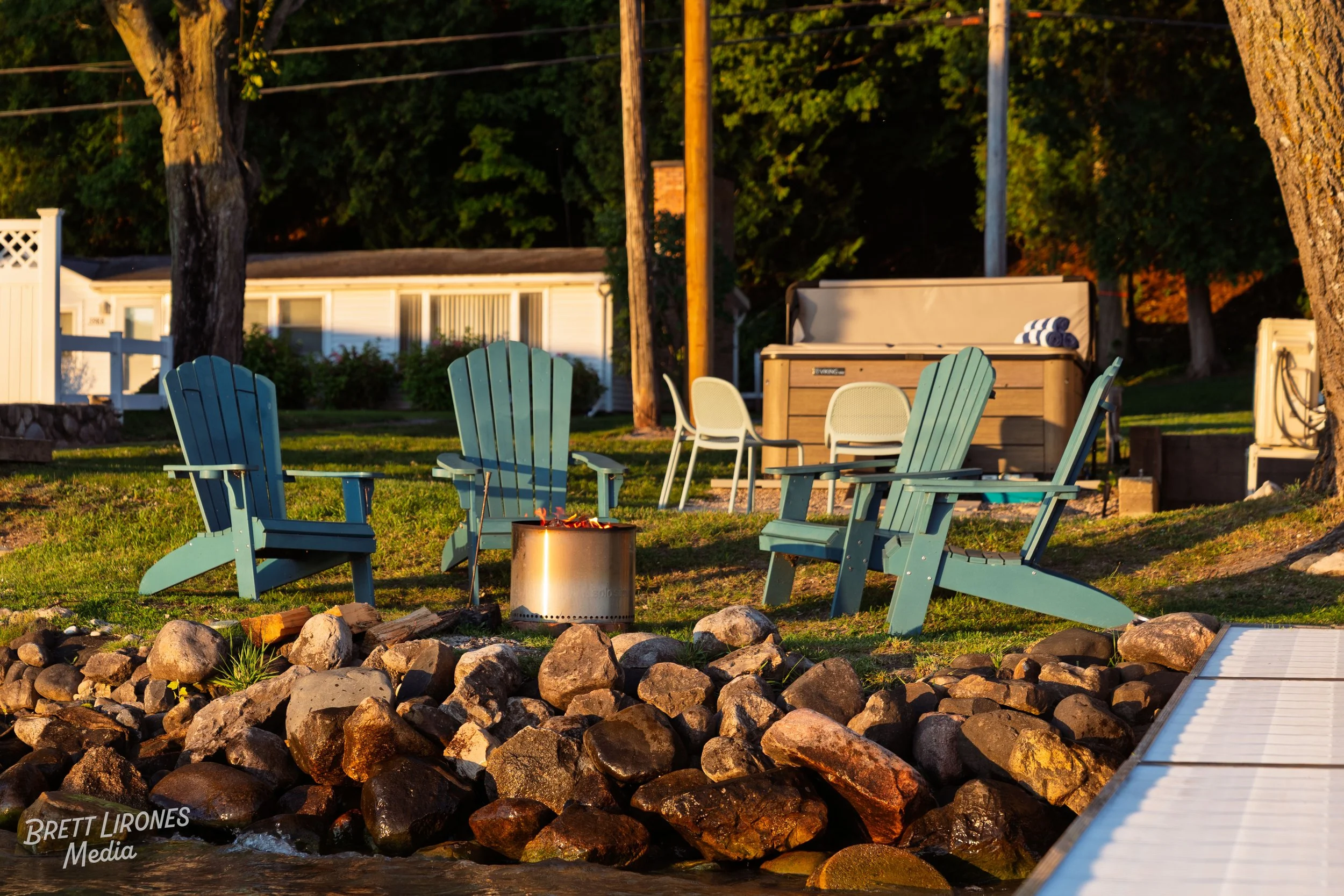 Four blue Adirondack chairs and two white side chairs arranged around a firepit on a grassy yard near a lake, with rocks lining the water's edge and a wooden hot tub in the background, surrounded by trees.