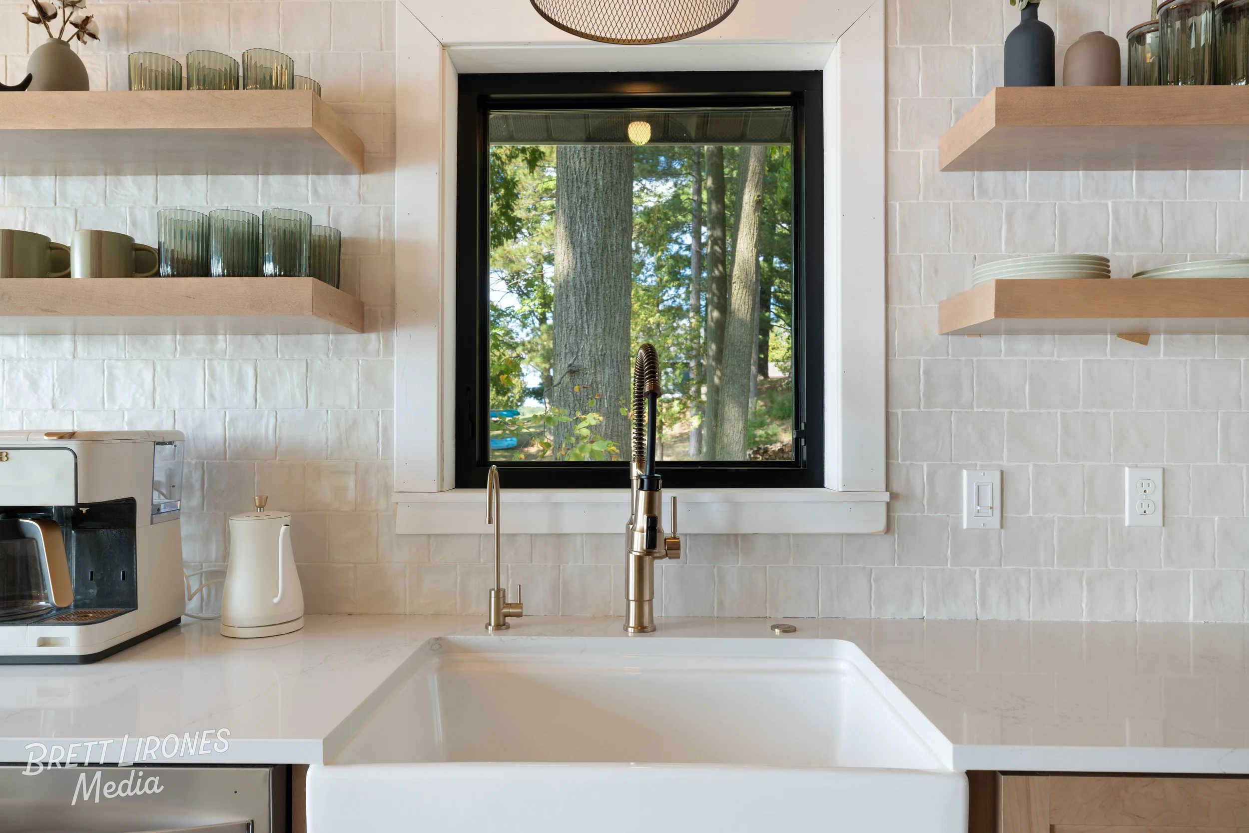 Kitchen with white brick backsplash, open wooden shelves with glassware and dishes, black-framed window showing trees outside, white countertop with sink, faucet, and small appliances.
