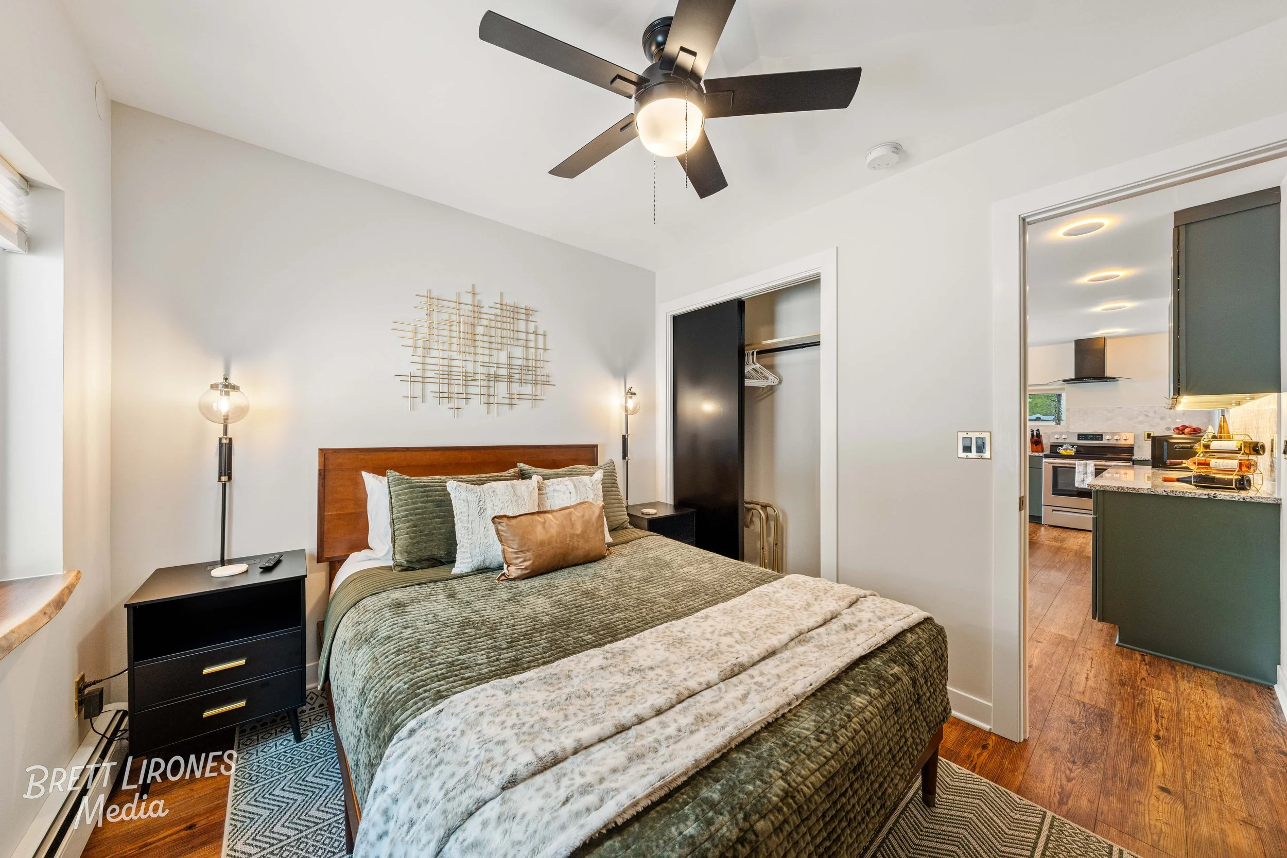 A bedroom with a wooden bed frame, green bedspread, multiple pillows, nightstands with lamps, a closet with sliding doors, and a view into a kitchen with countertops and appliances.