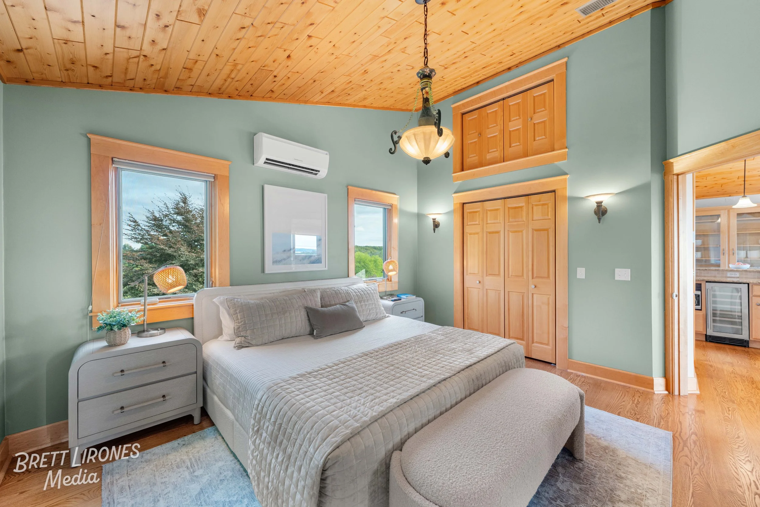 A bedroom with green walls, wooden ceiling, two windows, a bed with beige bedding, matching nightstands, small lamps, a pendant light, and a wooden closet door.