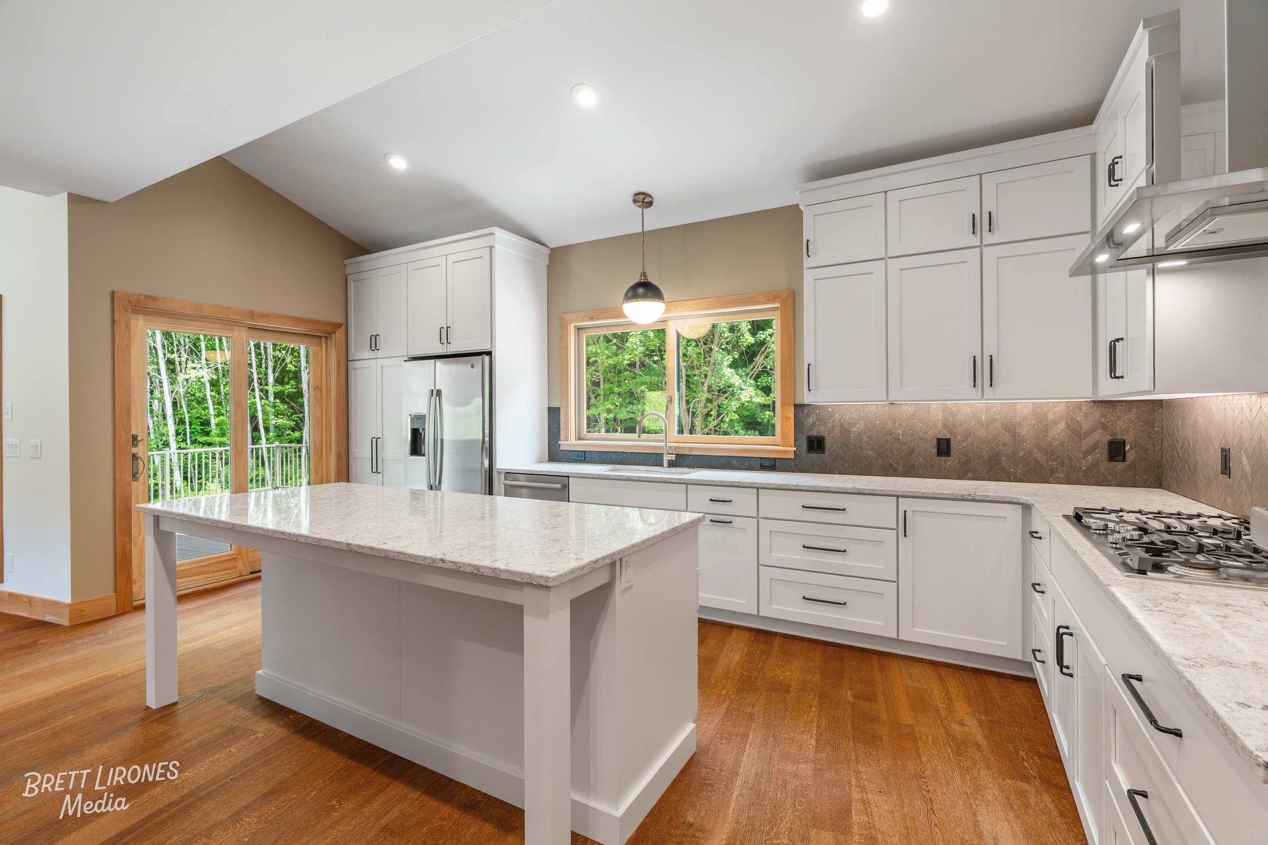 Modern kitchen with white cabinets, granite countertops, a central island, a stainless steel refrigerator, a window overlooking a wooded backyard, sliding glass doors, and hardwood floors.