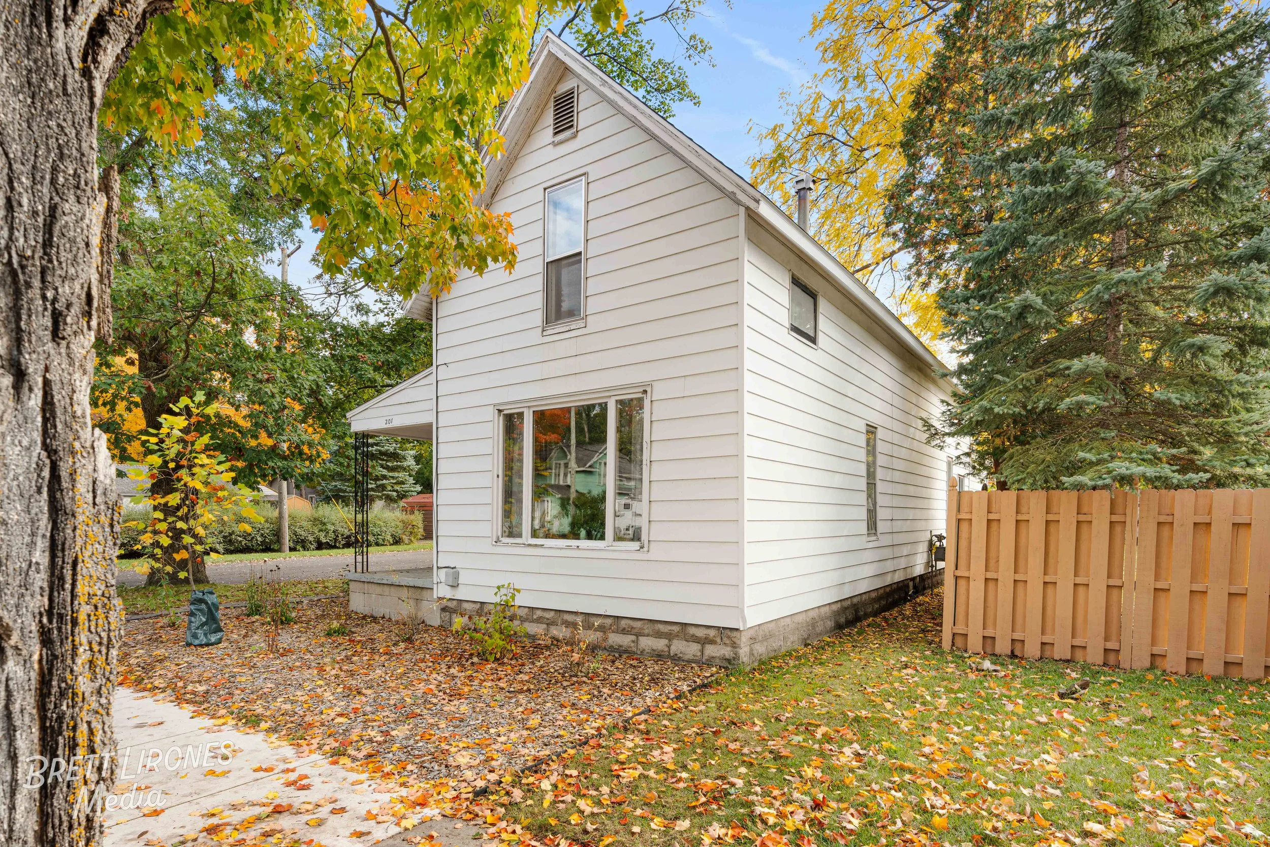 A two-story white house with a concrete foundation, large front window, surrounded by autumn trees with orange, yellow, and green leaves, and a wooden fence on the right side.
