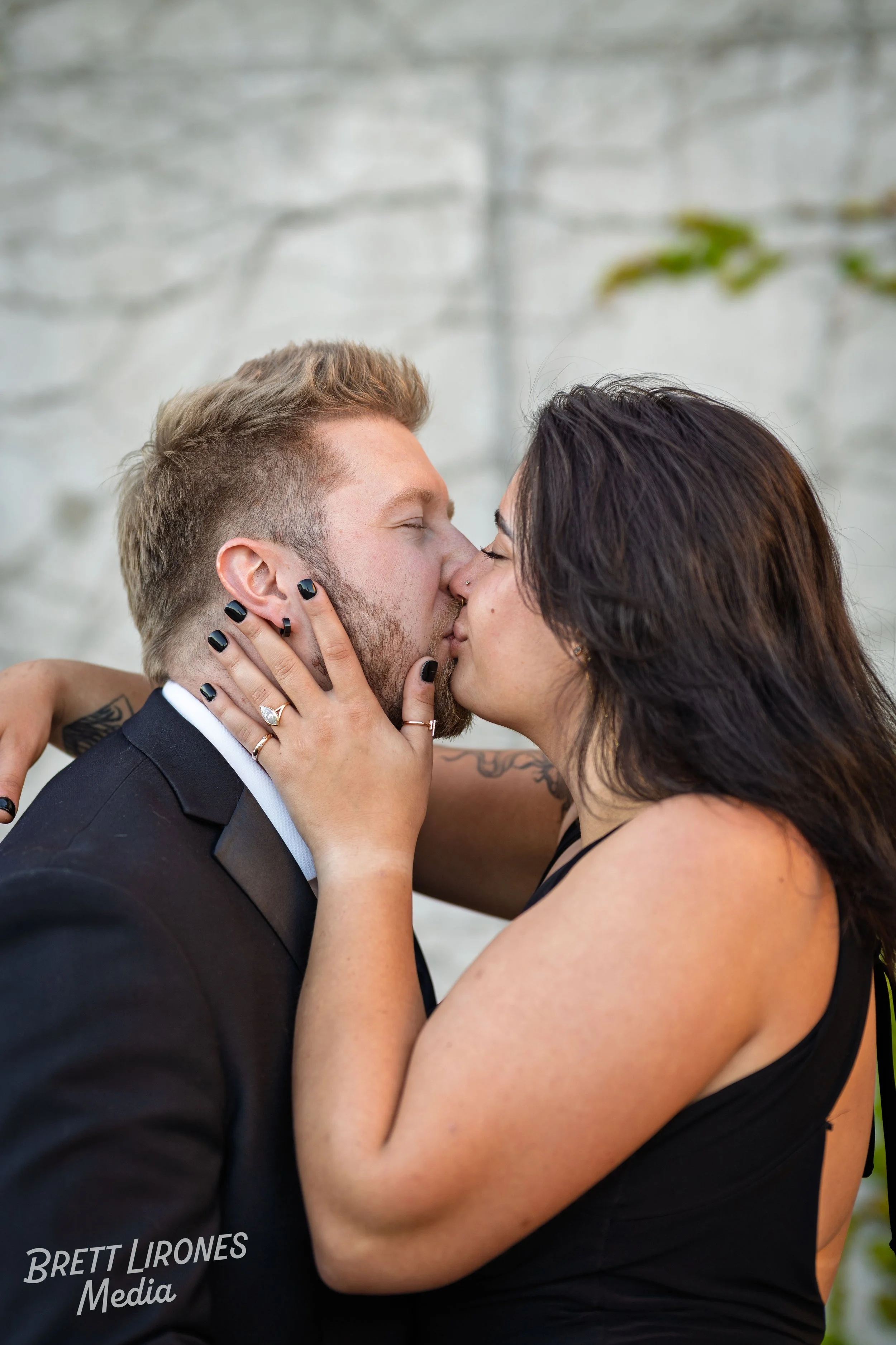 A couple sharing a kiss, the woman holding the man's face with her hands, both with closed eyes. The man wears a black tuxedo, and the woman wears a black sleeveless dress. The background is a blurred light-colored wall.