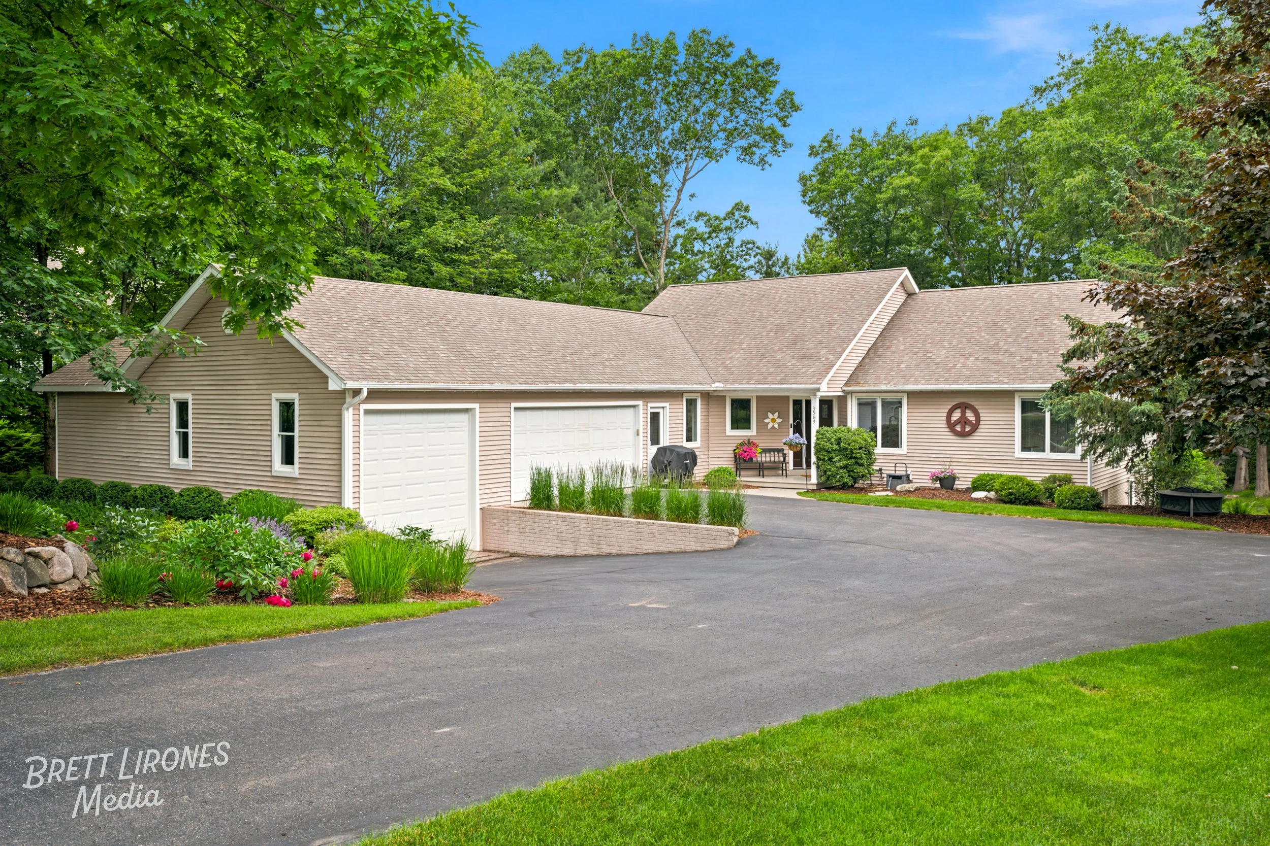 A single-story house with beige vinyl siding, a gabled roof, white garage door, and a front porch with potted flowers. The house is surrounded by lush green trees and a well-maintained lawn, with a curved driveway in front.