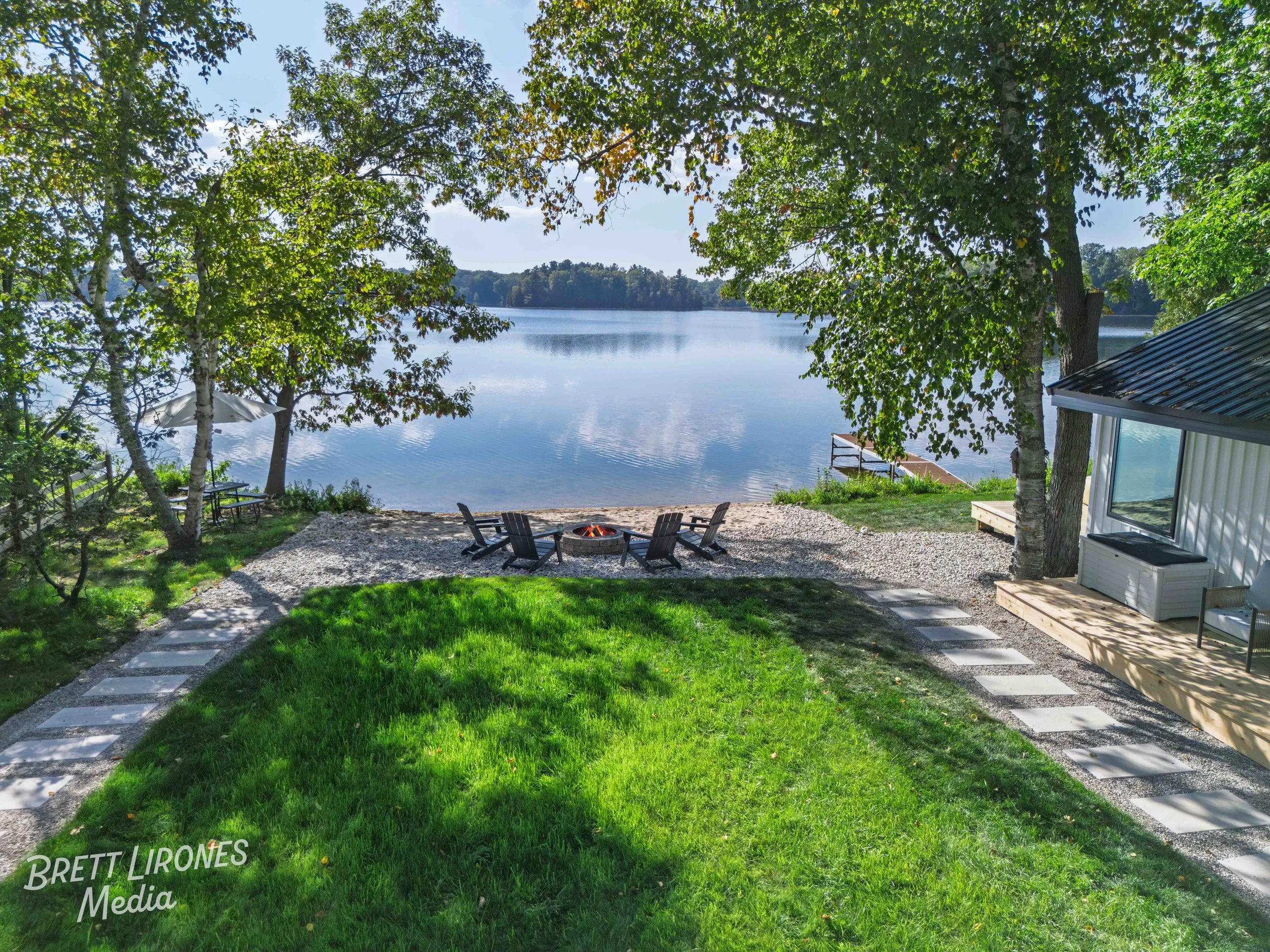 A lakeside scene featuring a grassy yard with a circular fire pit surrounded by four chairs, tall trees providing shade, and a wooden dock extending into the calm lake. There is a house with a metal roof on the right, and a gravel pathway leading to 