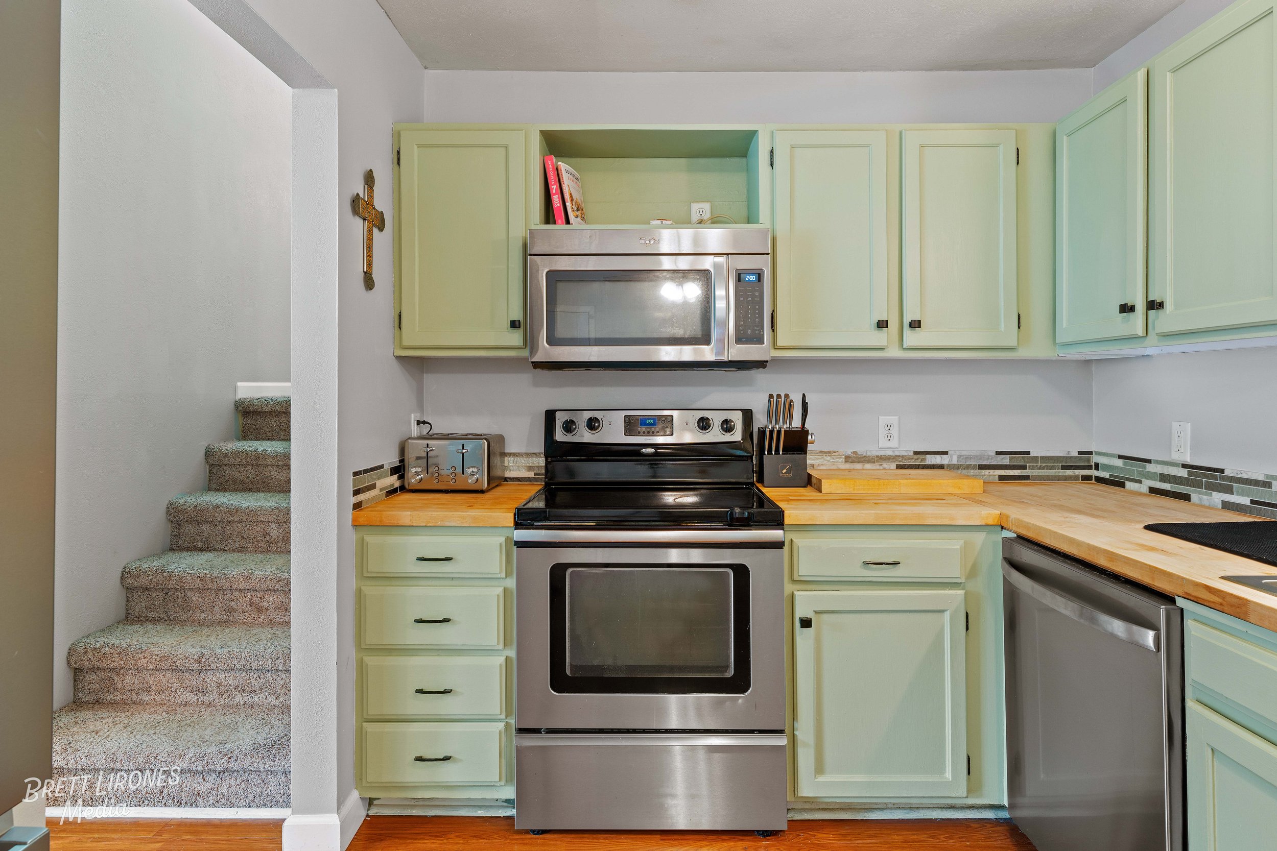 Kitchen with light green cabinets, stainless steel stove and microwave, wooden countertops, toaster, knife block, and a staircase with carpeted steps to the left.