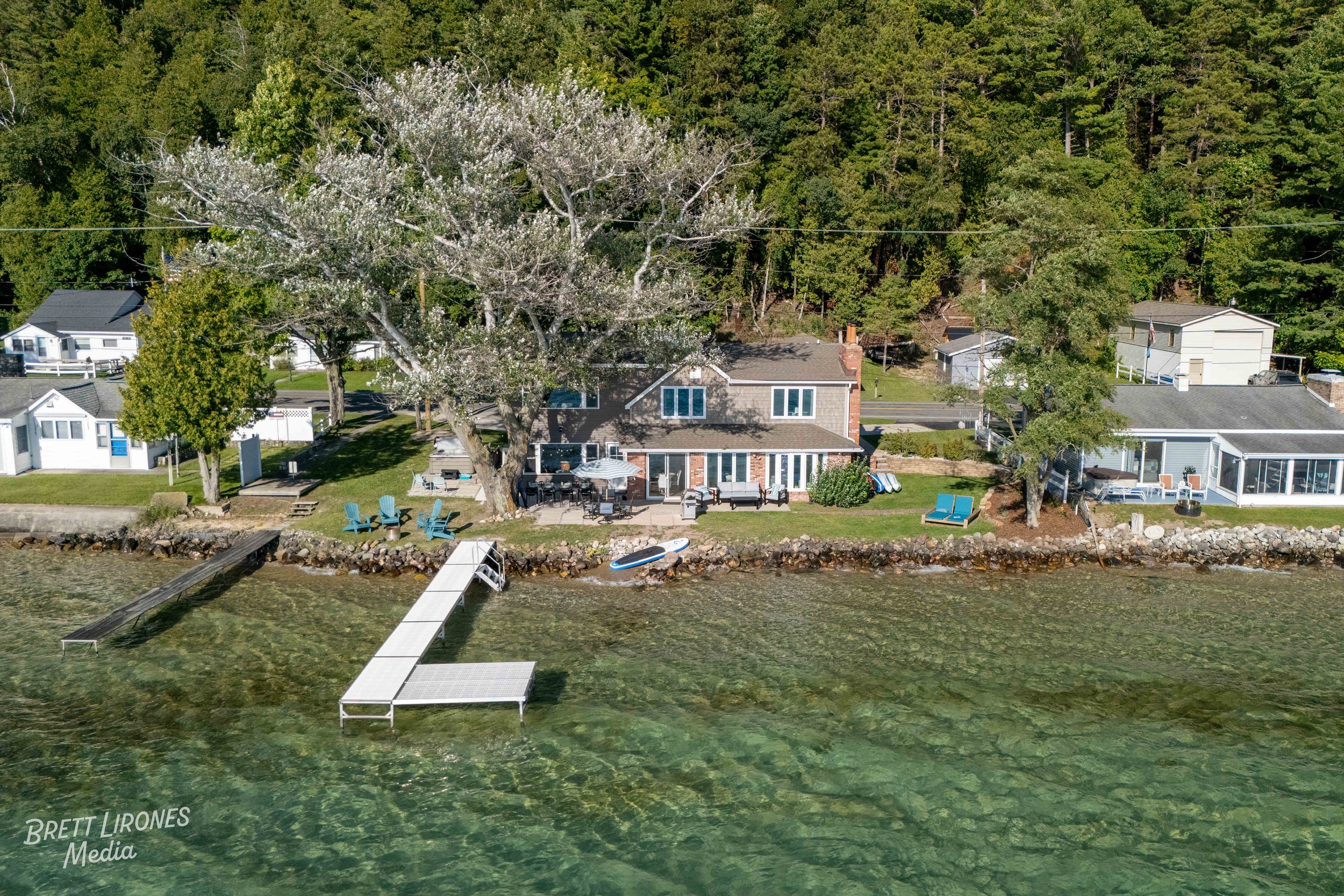 A lakeside house with a backyard featuring blue Adirondack chairs, a paddleboard, a dock, and outdoor furniture, surrounded by trees and neighboring houses.