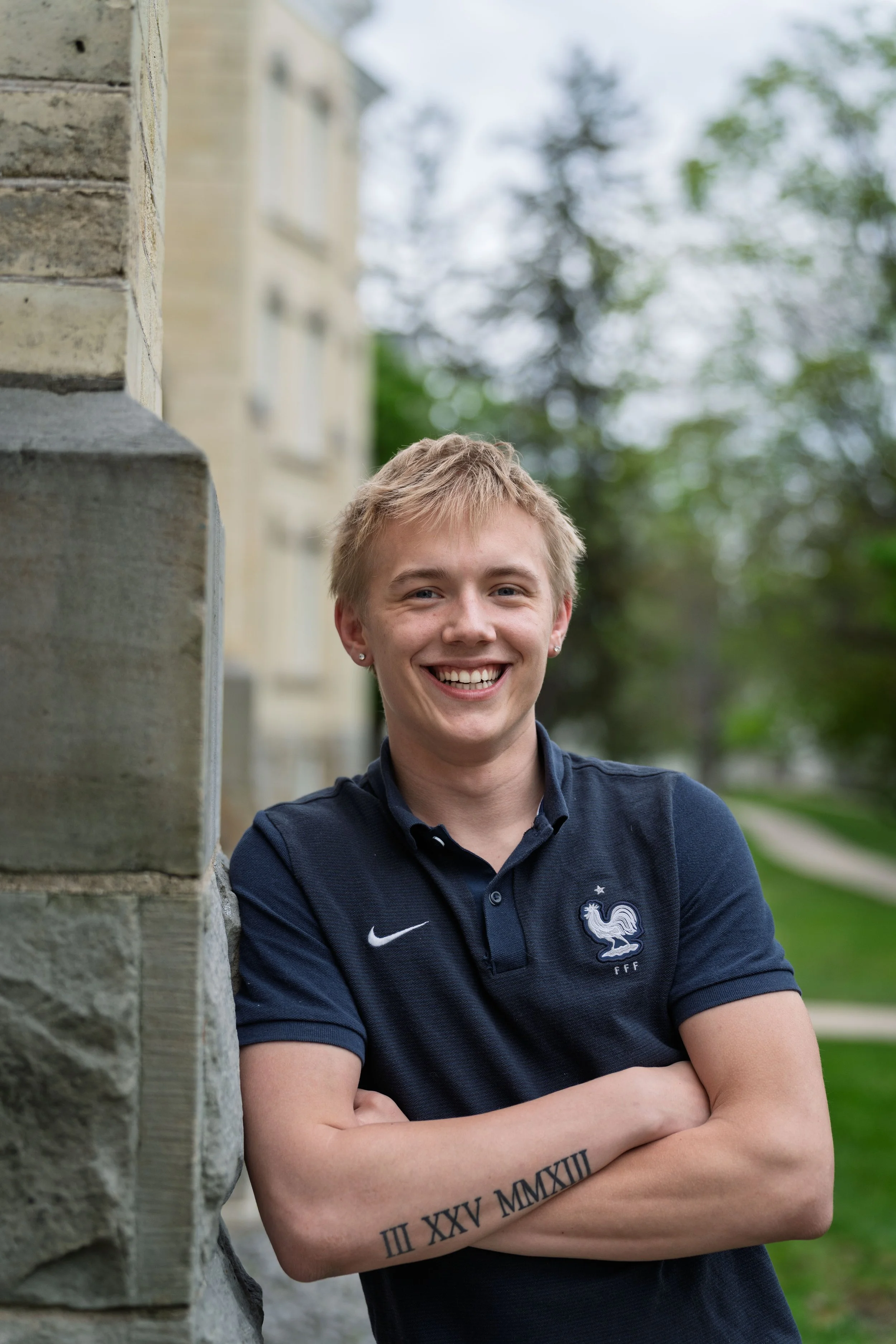 A young person with short blonde hair, wearing a navy blue polo shirt with a French football emblem, smiling and standing outdoors with crossed arms on a cloudy day.