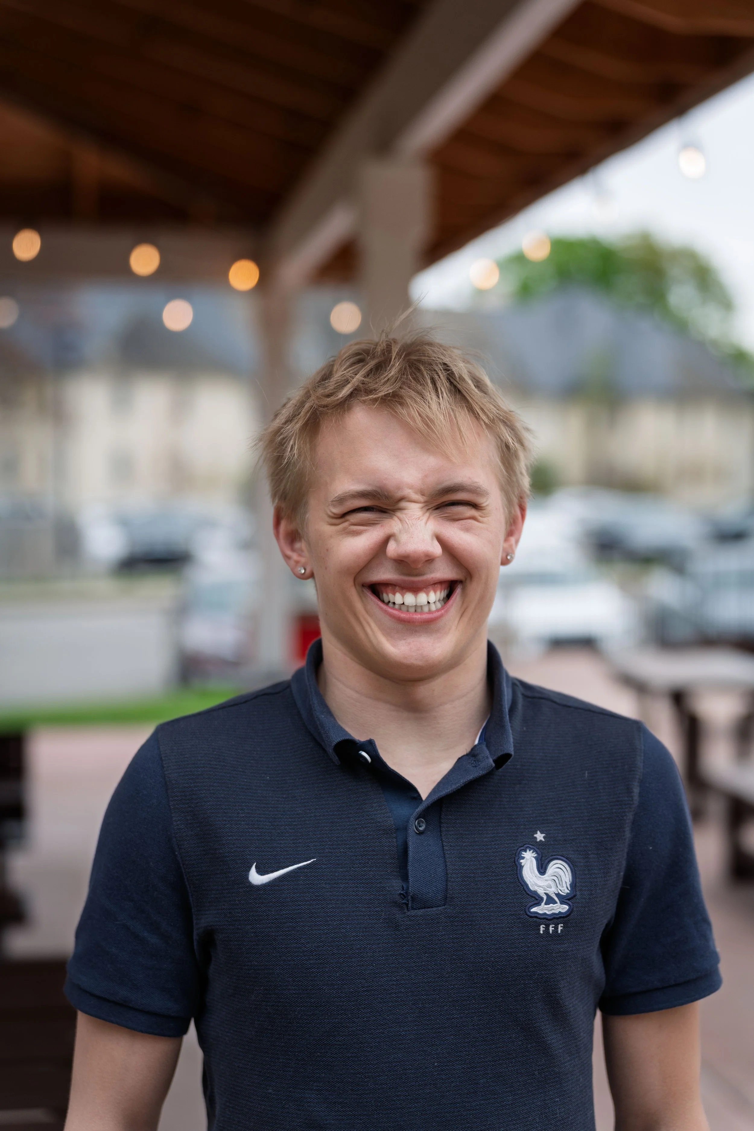 A young woman with short blonde hair, wearing a navy blue polo shirt with a French football team's emblem, smiling with eyes closed in an outdoor setting with string lights and a blurred background of buildings and parked cars.