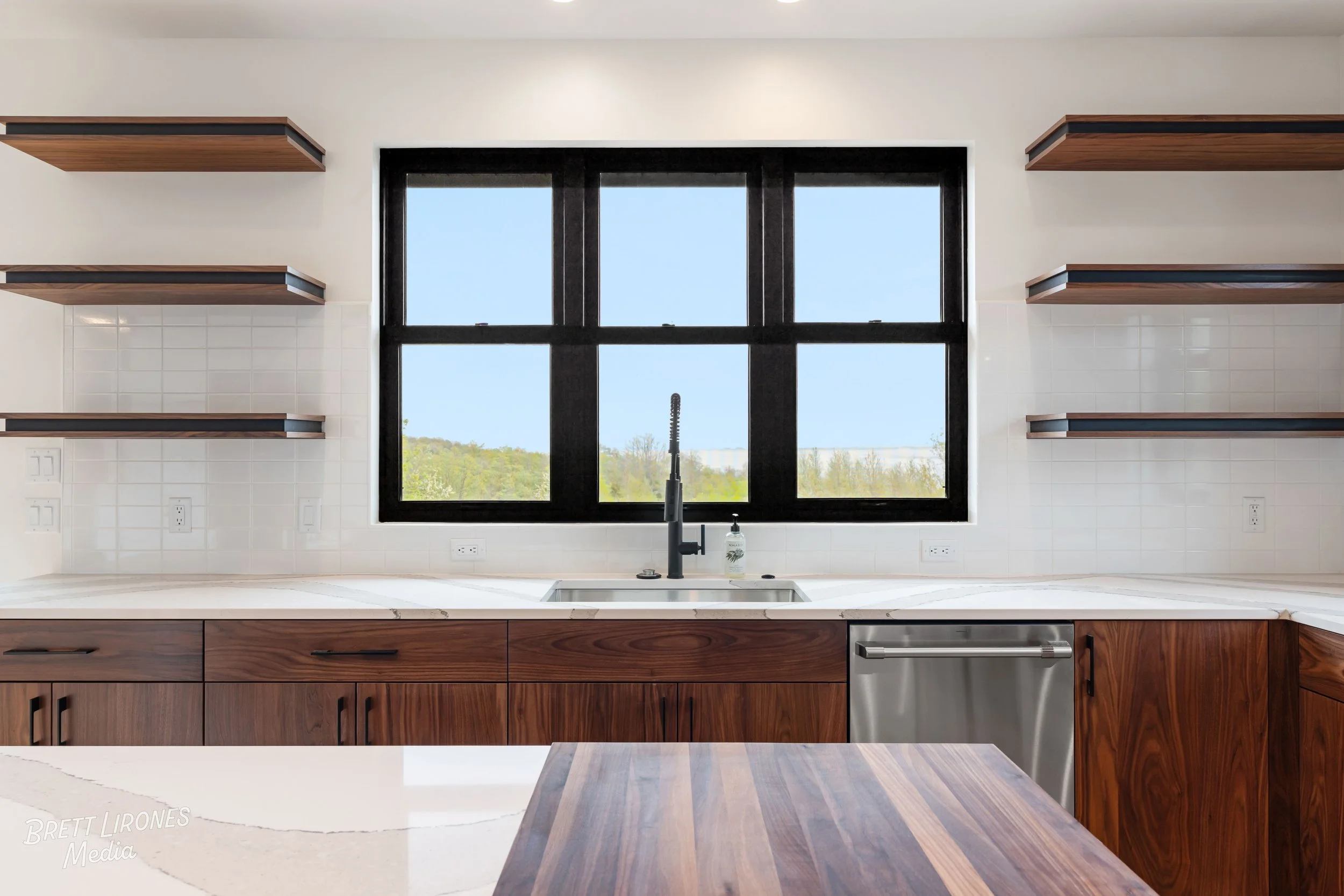Modern kitchen with a large black-framed window above a white marble countertop, featuring a stainless steel dishwasher and wooden cabinets, with open wooden shelves on white tiled walls.