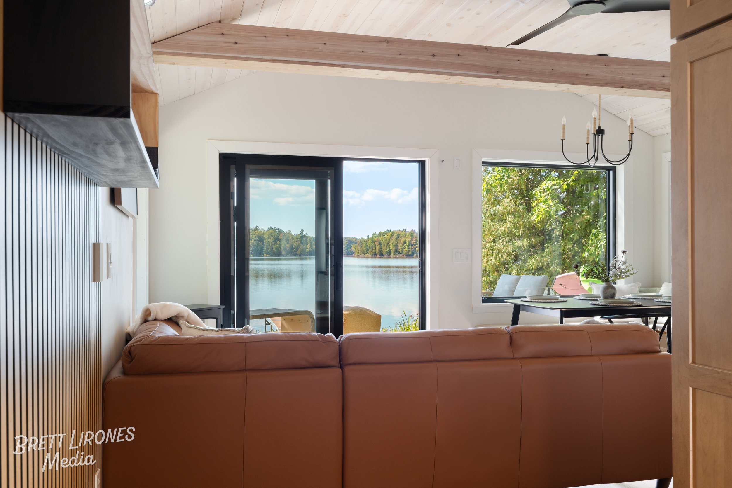 View from inside a living room looking out to a lake through sliding glass door and window, with a brown sofa in the foreground, and a dining table with a chandelier hanging above.