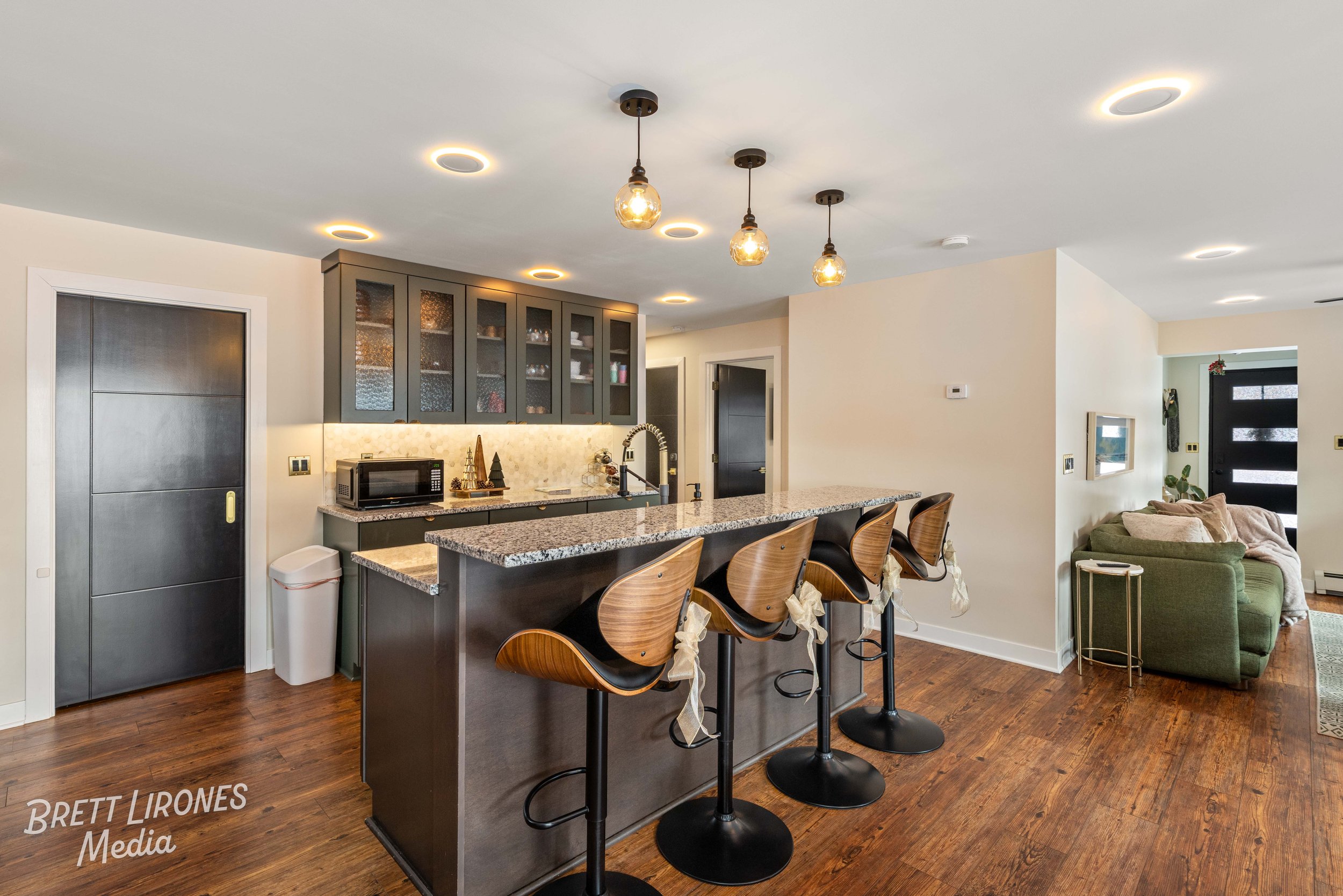 Modern kitchen with black cabinets, granite countertop island, three barstools with wooden backs, pendant lights, and a cozy living area with a green sofa.