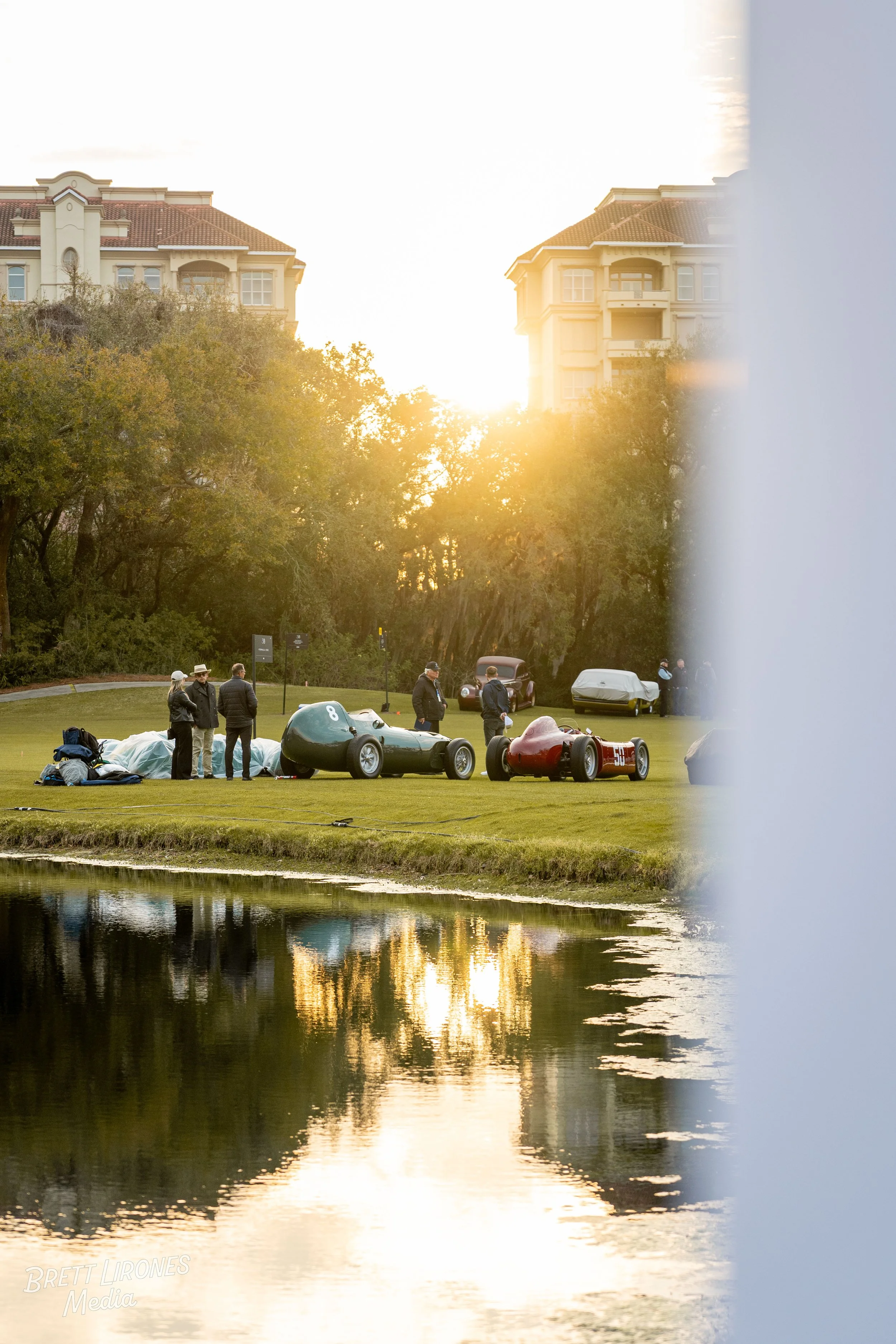 A group of people preparing a vintage race car on a grassy area by a pond during a sunset, with large buildings in the background.
