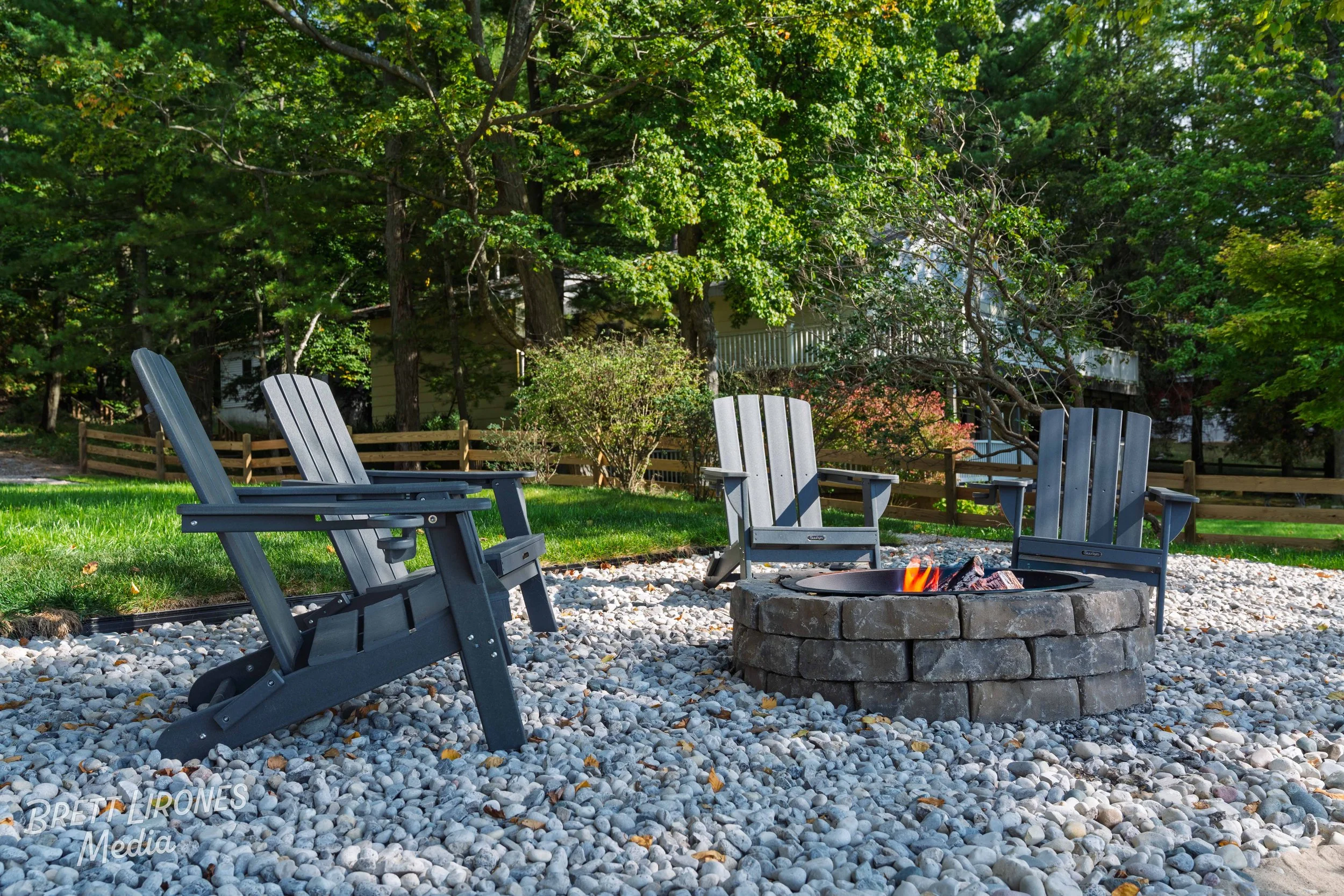 Four Adirondack chairs arranged around a fire pit on a stone-paved area, with a backdrop of green trees and a wooden fence.