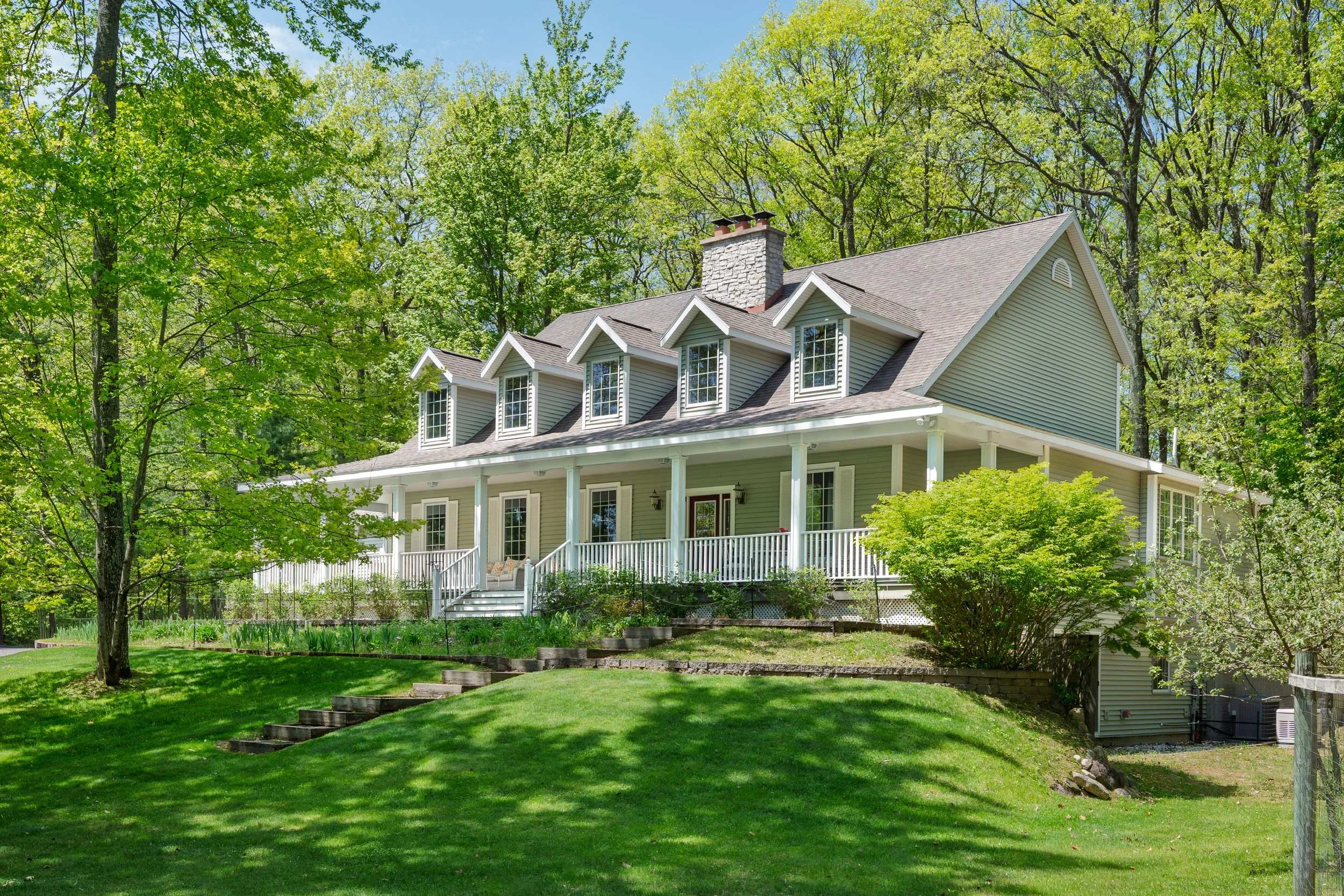 A large house with four dormer windows on the upper floor, a porch with white railings, set in a lush green backyard with trees and a well-maintained lawn.