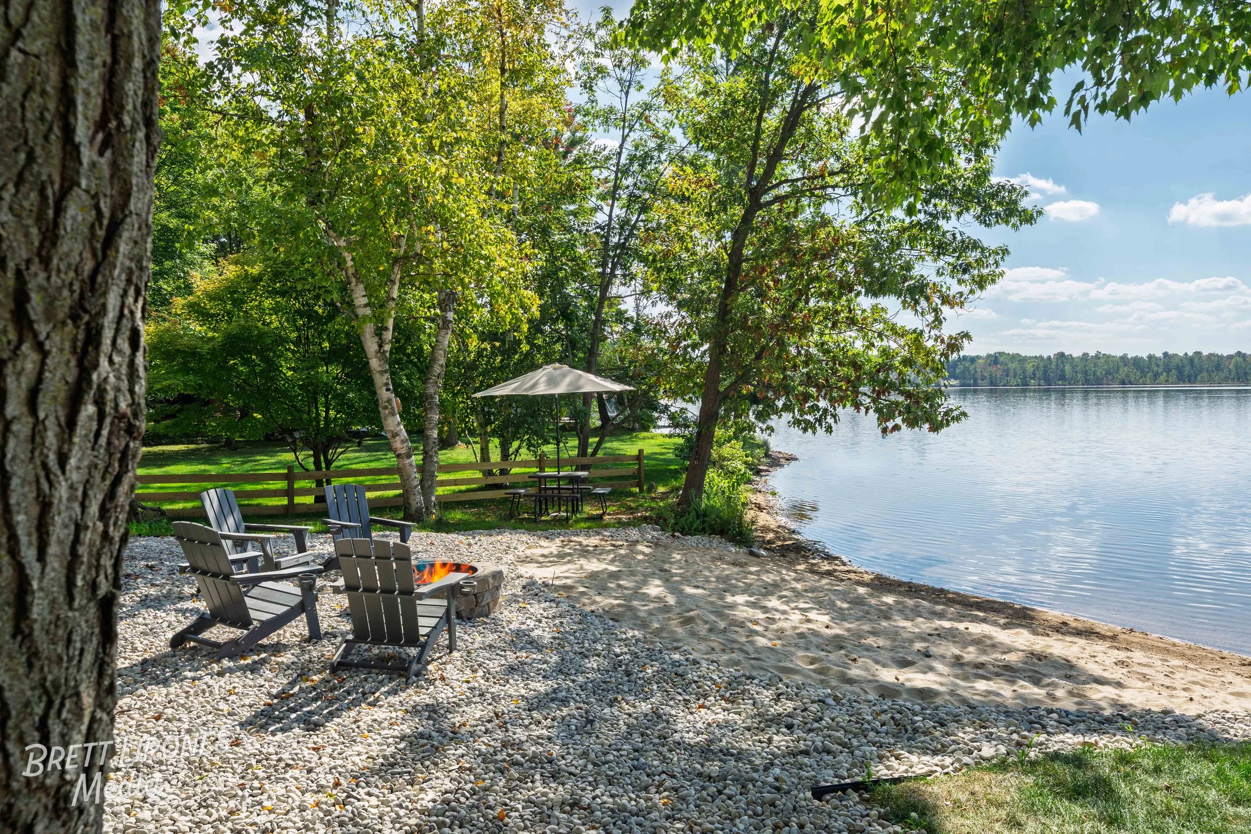 A lakeside outdoor scene with adirondack chairs around a fire pit, a shaded picnic table under an umbrella, green trees, and a sandy shoreline extending to a calm lake under a partly cloudy sky.