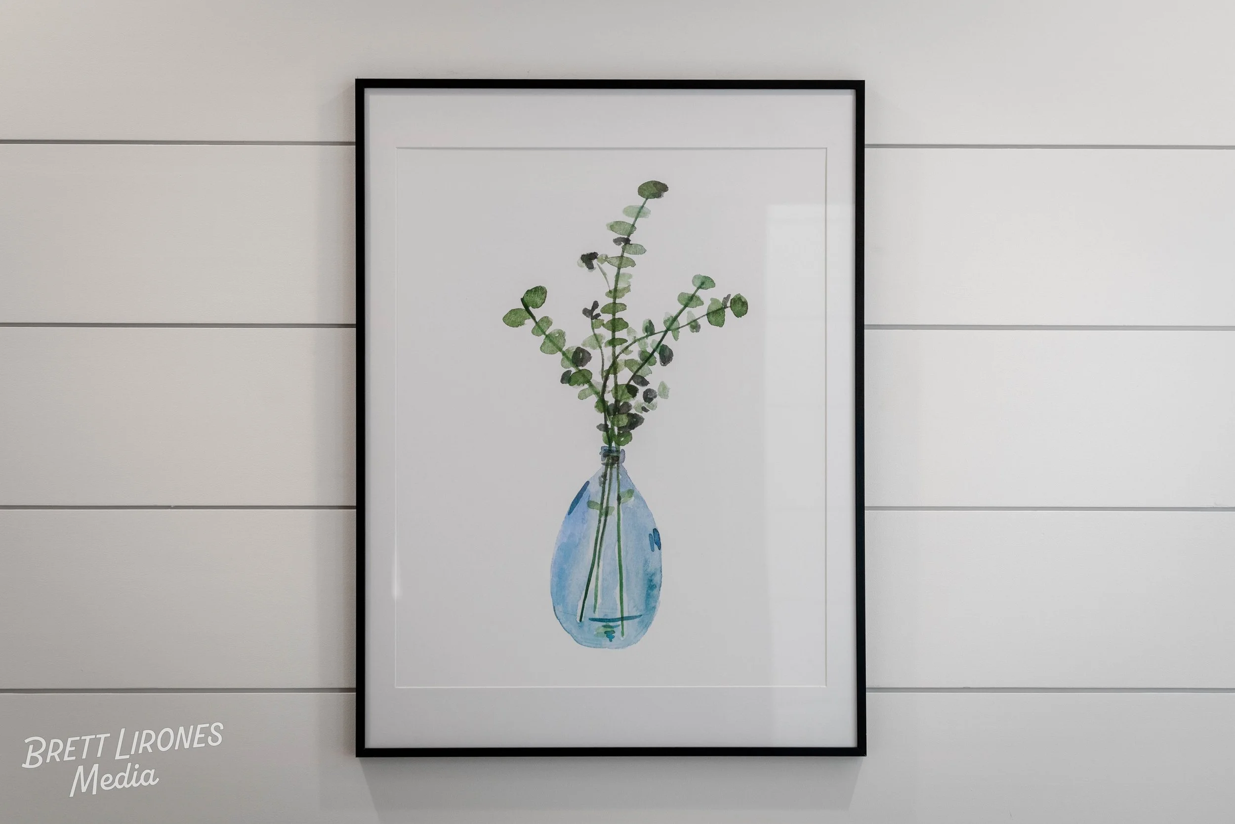 A framed watercolor painting of a green leafy plant in a blue vase, hanging on a white wall with horizontal panels.
