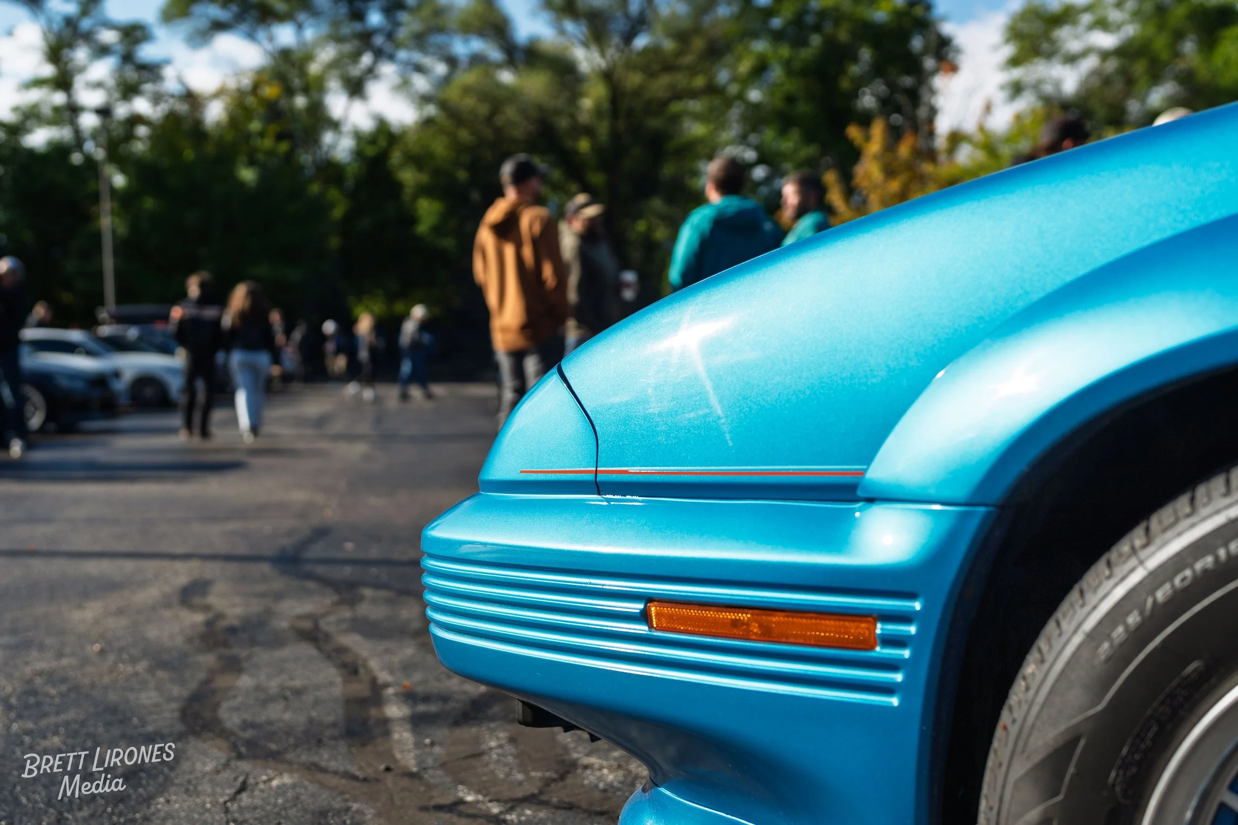 Close-up of the front part of a vintage blue sports car with a group of people gathered in the background in a parking lot surrounded by trees.