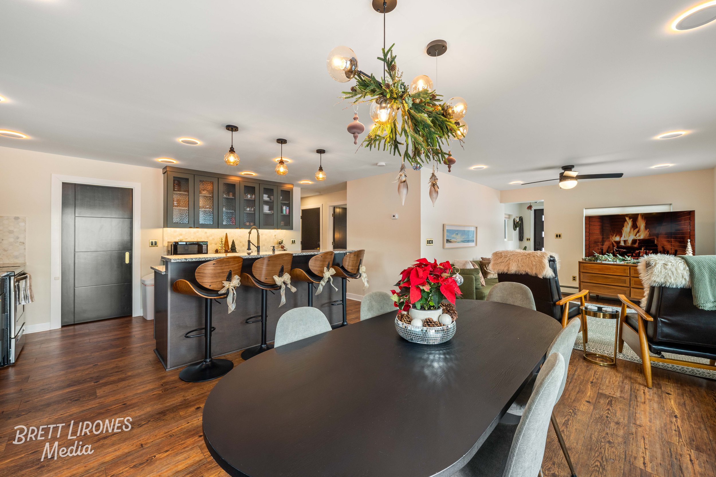 Open-concept living and dining area decorated for Christmas with a hanging botanical chandelier, poinsettia centerpiece, and fireplace with a fire, holiday decor, and plush furniture.