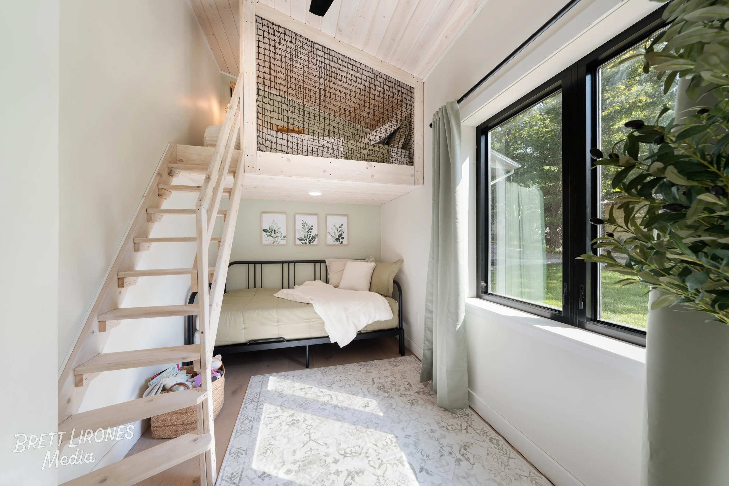 A cozy bedroom with a black metal bed frame, light-colored bedding, and decorative botanical art hanging above. A large window with light green curtains lets in natural sunlight. Above the bed, there's a loft space enclosed with a wooden frame and bl