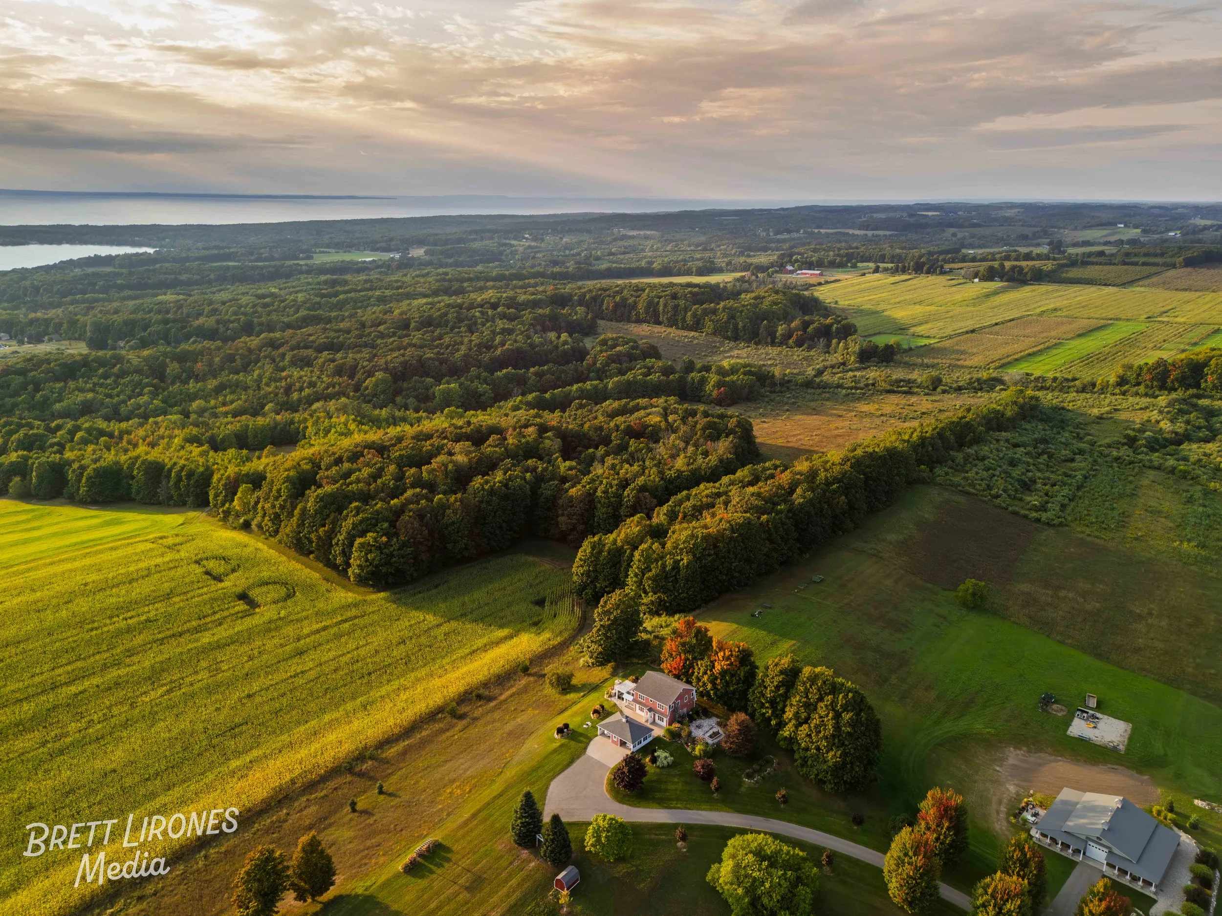 Aerial view of a rural landscape featuring a house, a garage, and open farmland with fields and trees, with a lake in the distance under a cloudy sky.