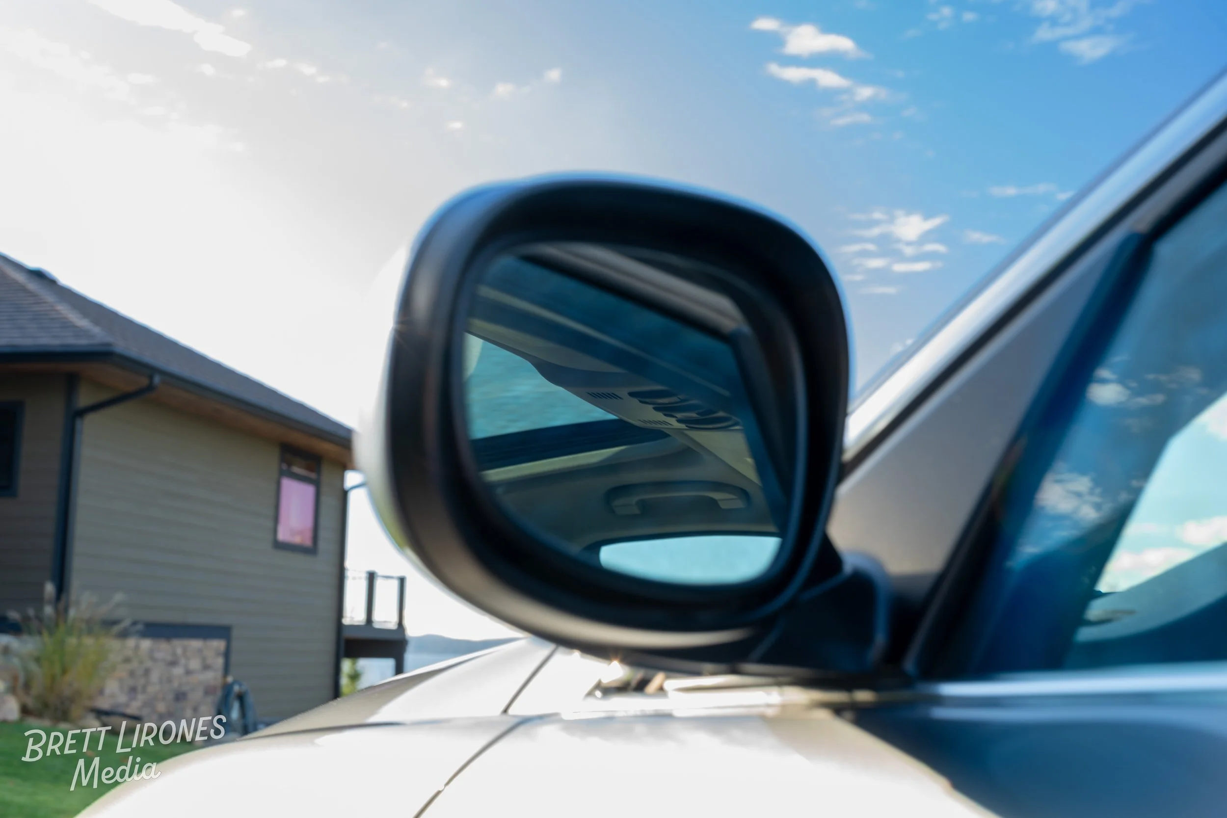 Close-up of a side mirror on a silver vehicle reflecting a blue sky with clouds, a house with a deck, and some greenery.