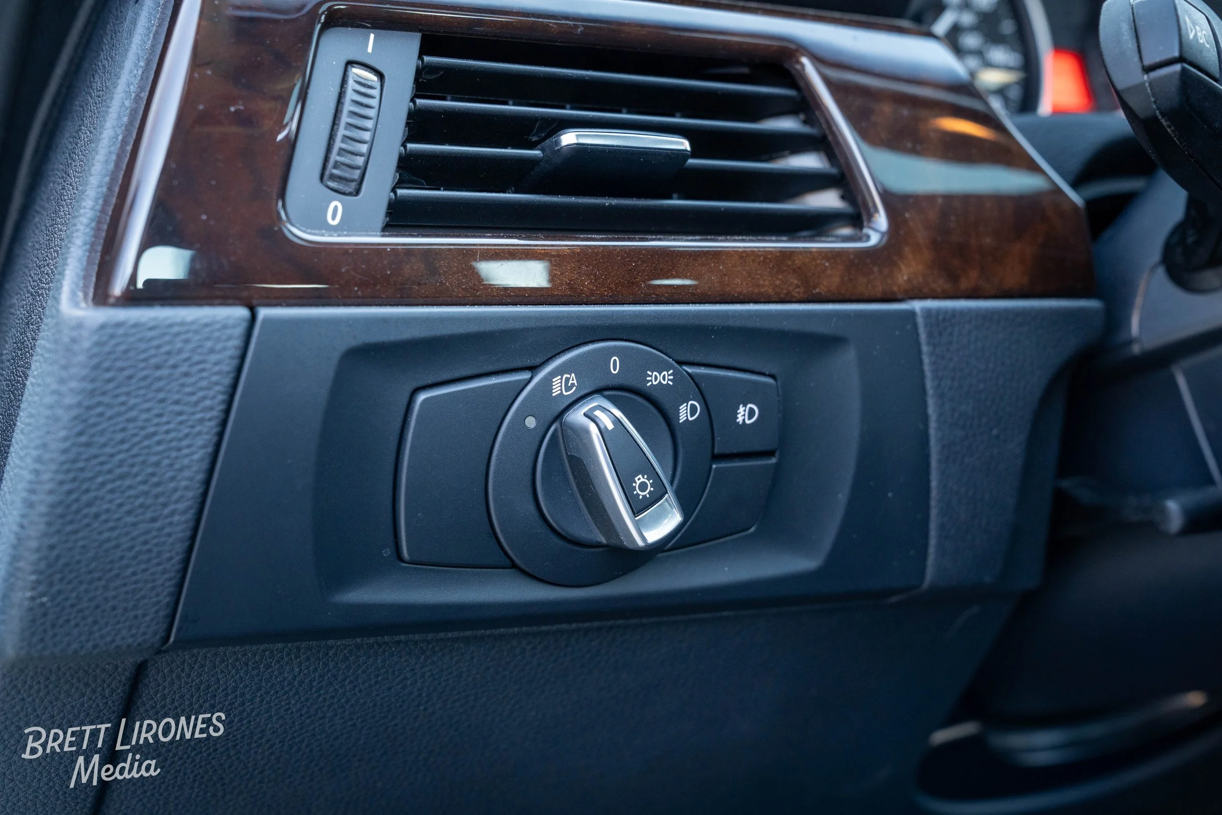 Close-up of a car's headlight control switch with multiple settings, located below a rectangular air vent with adjustable louvers, on a dashboard with wood grain trim.