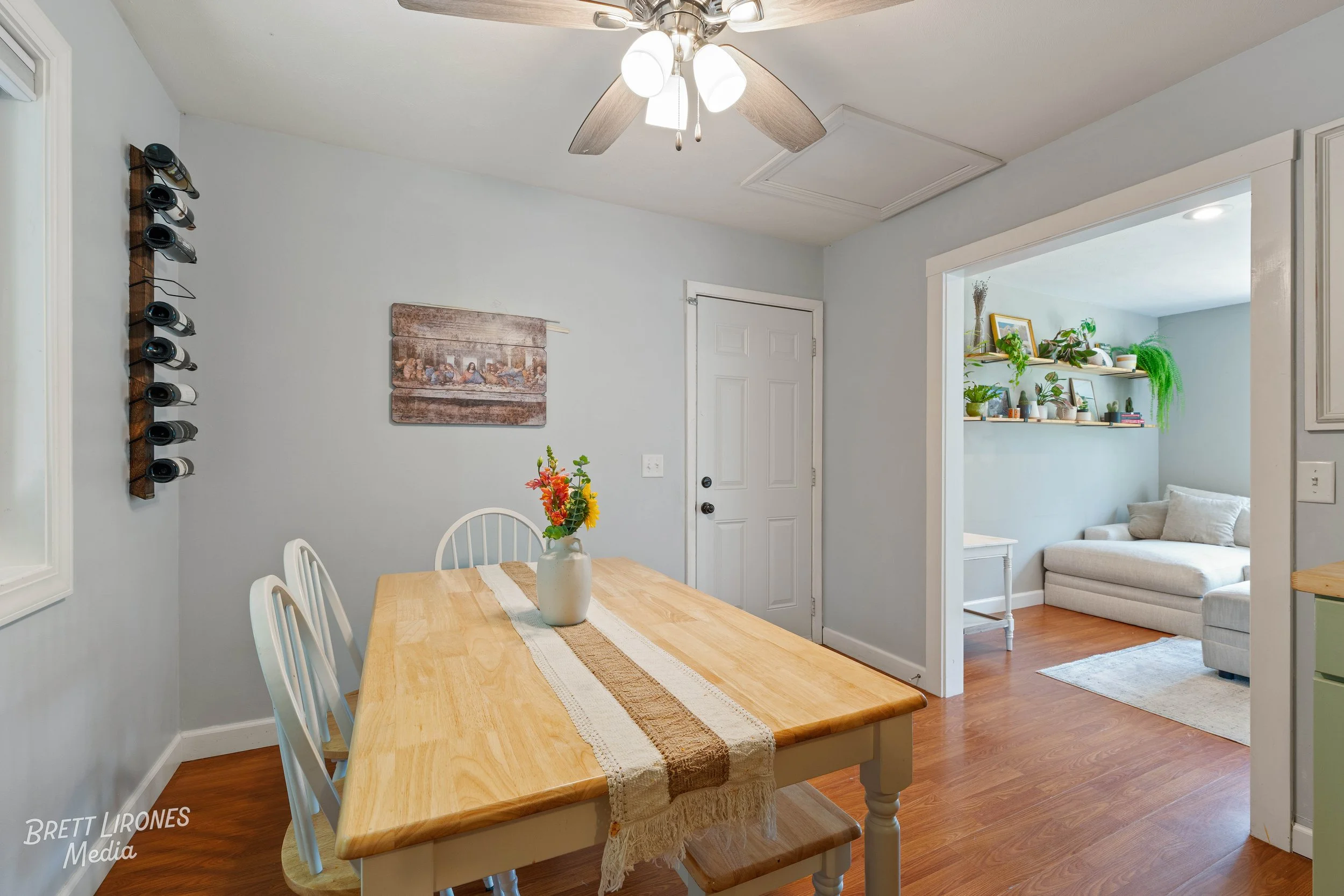 A dining room with a wooden table, four white chairs, and a vase with colorful flowers. Adjacent to a living room with a light-colored sofa, shelves with plants, and artwork. Ceiling fan with light fixtures. Light-colored walls and hardwood floors.