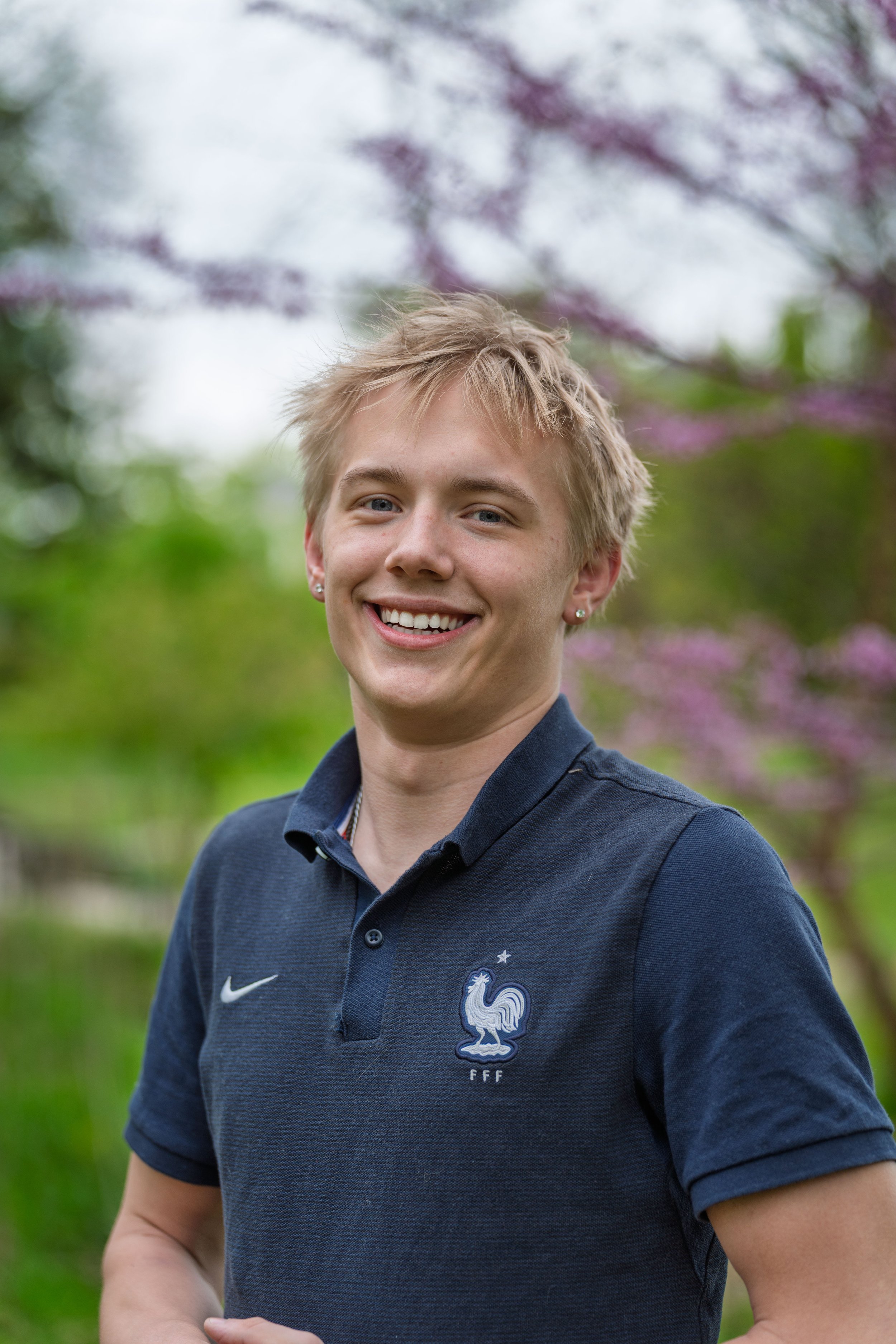 Young man with short blonde hair smiling, wearing a navy blue France national football team polo shirt, outdoors with blurred green trees and purple blossoms in the background.