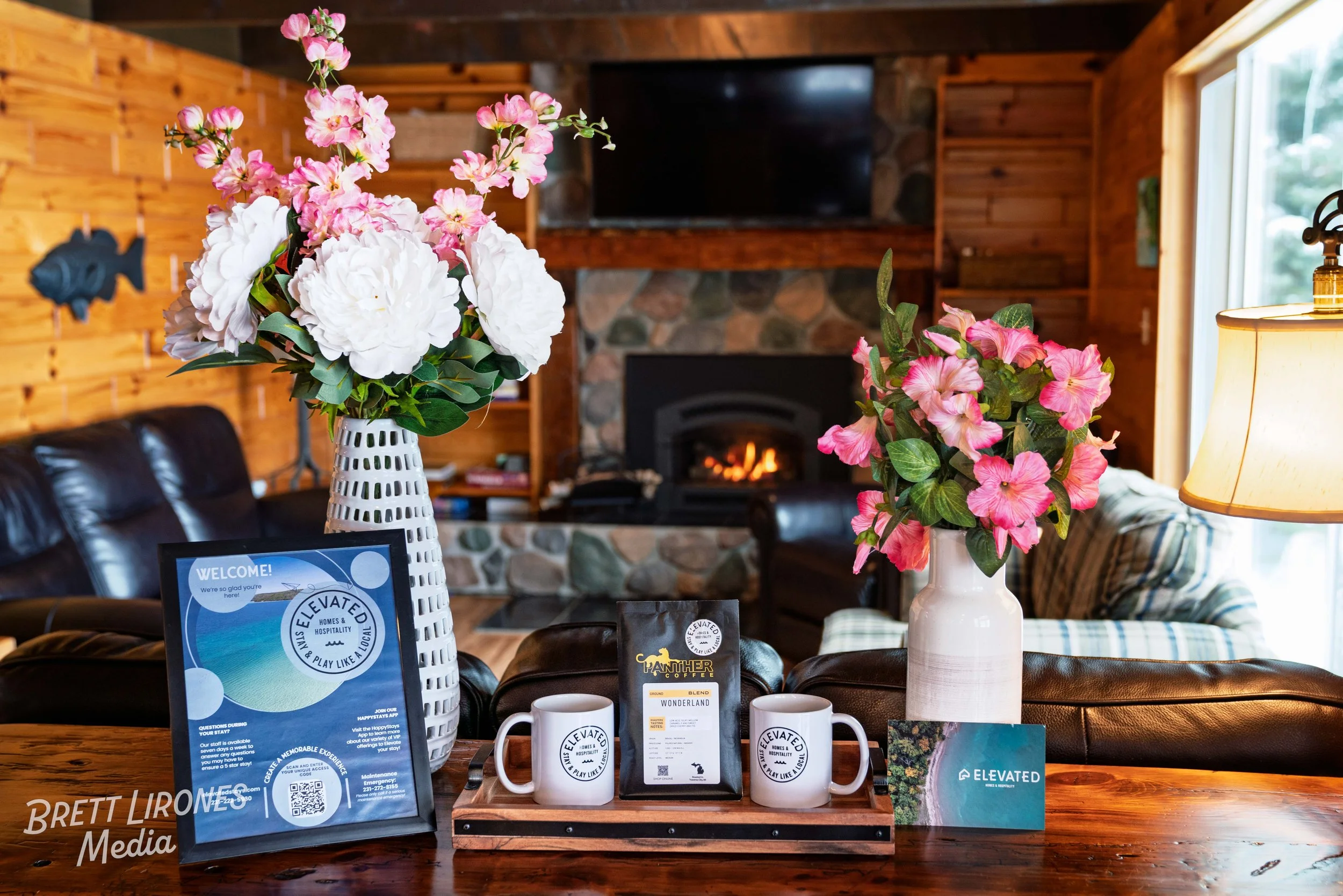 Decorative arrangement of pink and white flowers in vases on a wooden table, with promotional materials and coffee cups, inside a cozy wood-paneled living room with a stone fireplace and a mounted flat-screen TV.