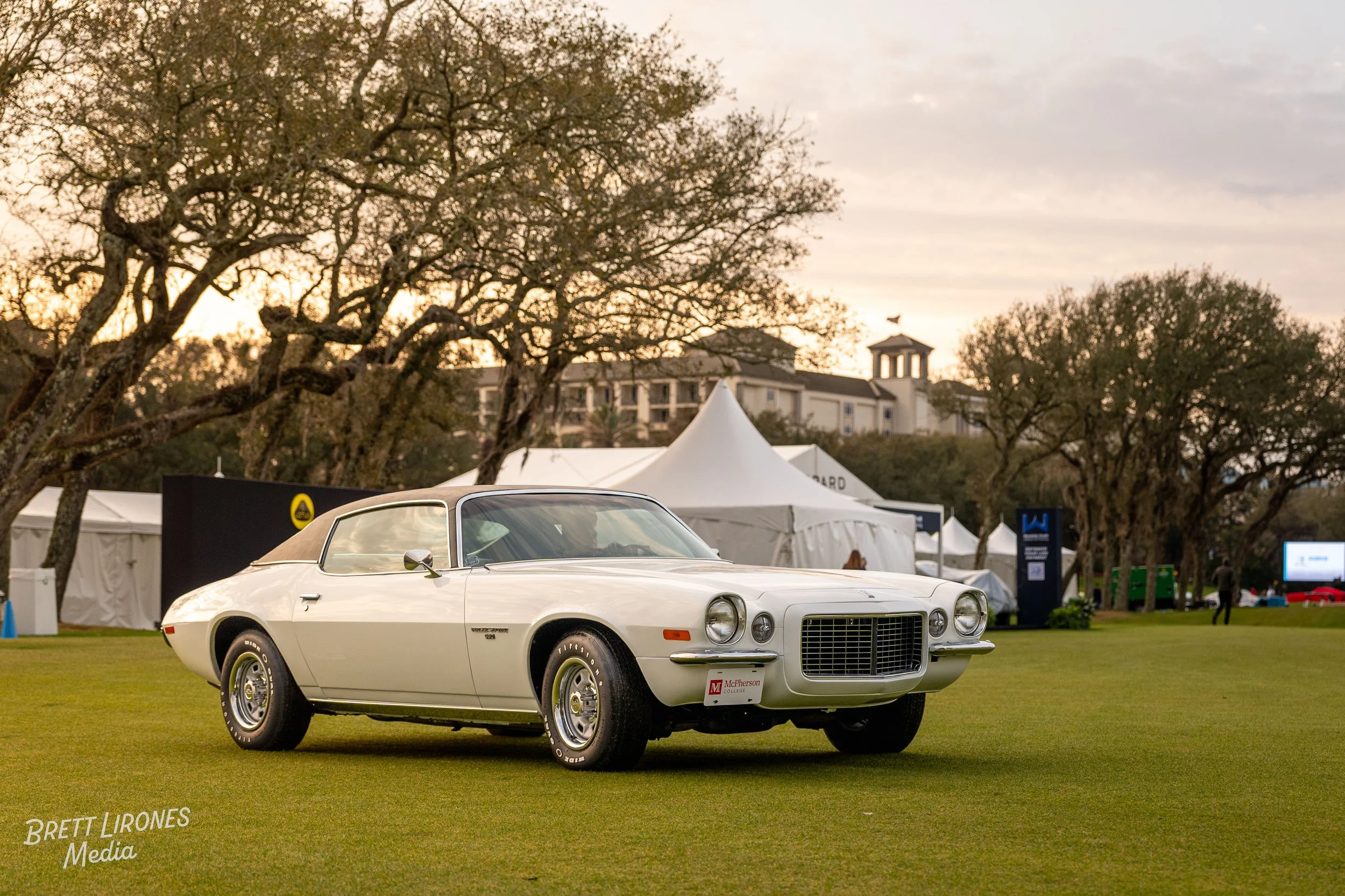 A classic white convertible car displayed on a grassy field at an outdoor event, with white tents and trees in the background during sunset.