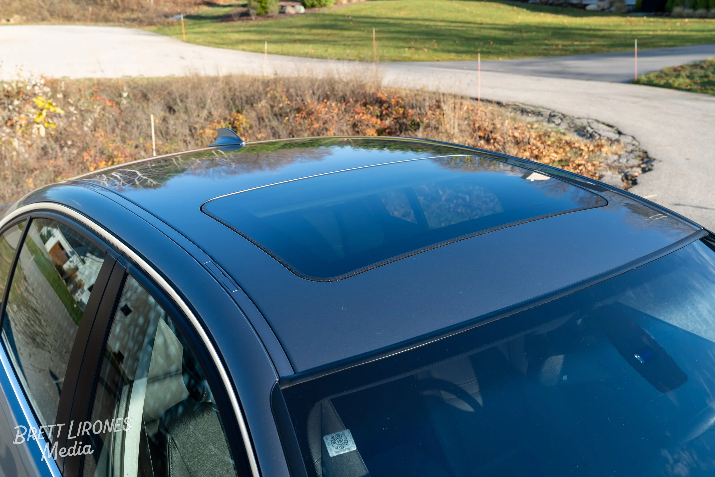 A black car with a sunroof parked on a suburban street with a curved driveway and trees in the background.