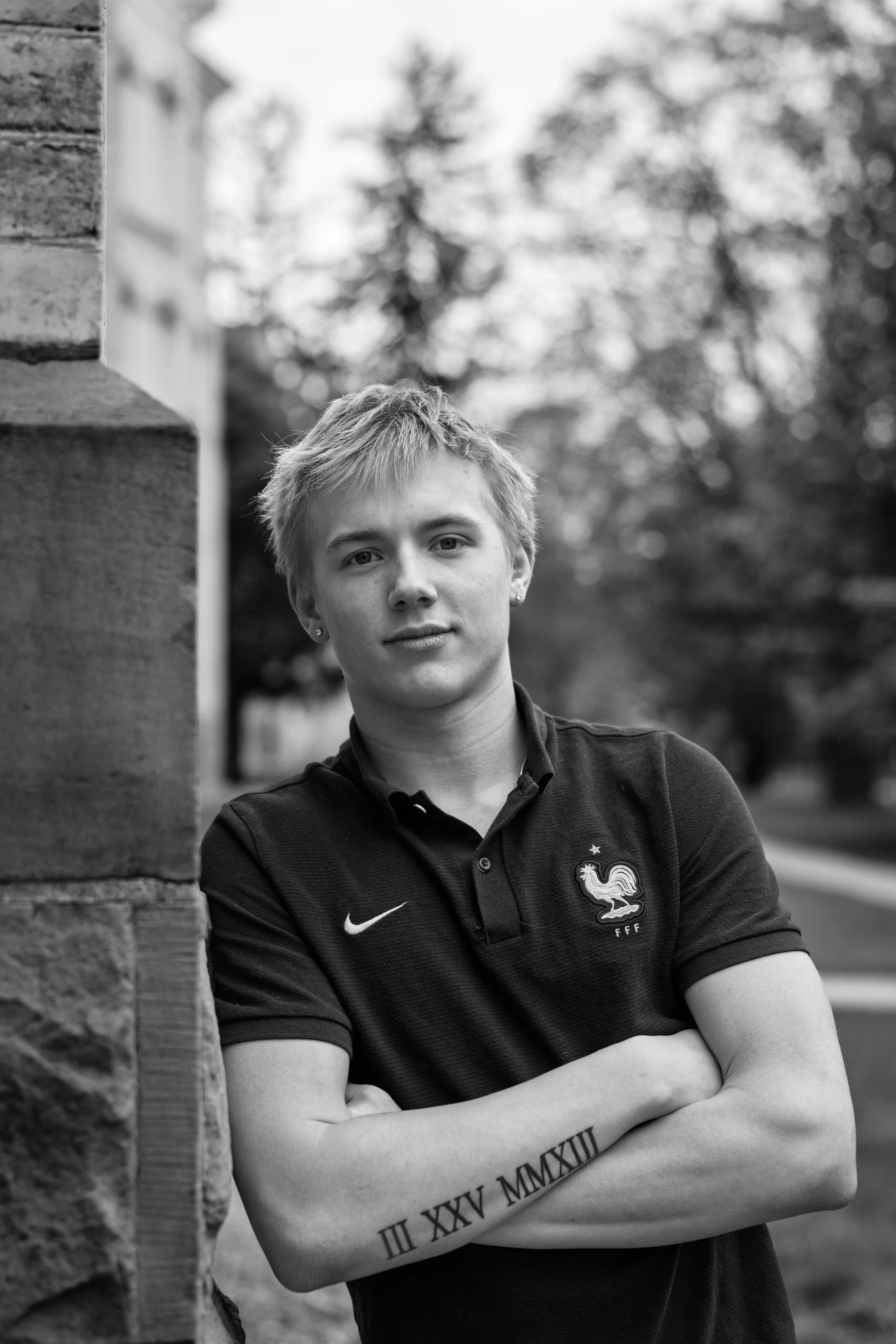 Black and white portrait of a young man with short blond hair, leaning against a brick wall outdoors, wearing a black polo shirt with French national team crest, arms crossed, with a tattoo of Roman numerals on his forearm.