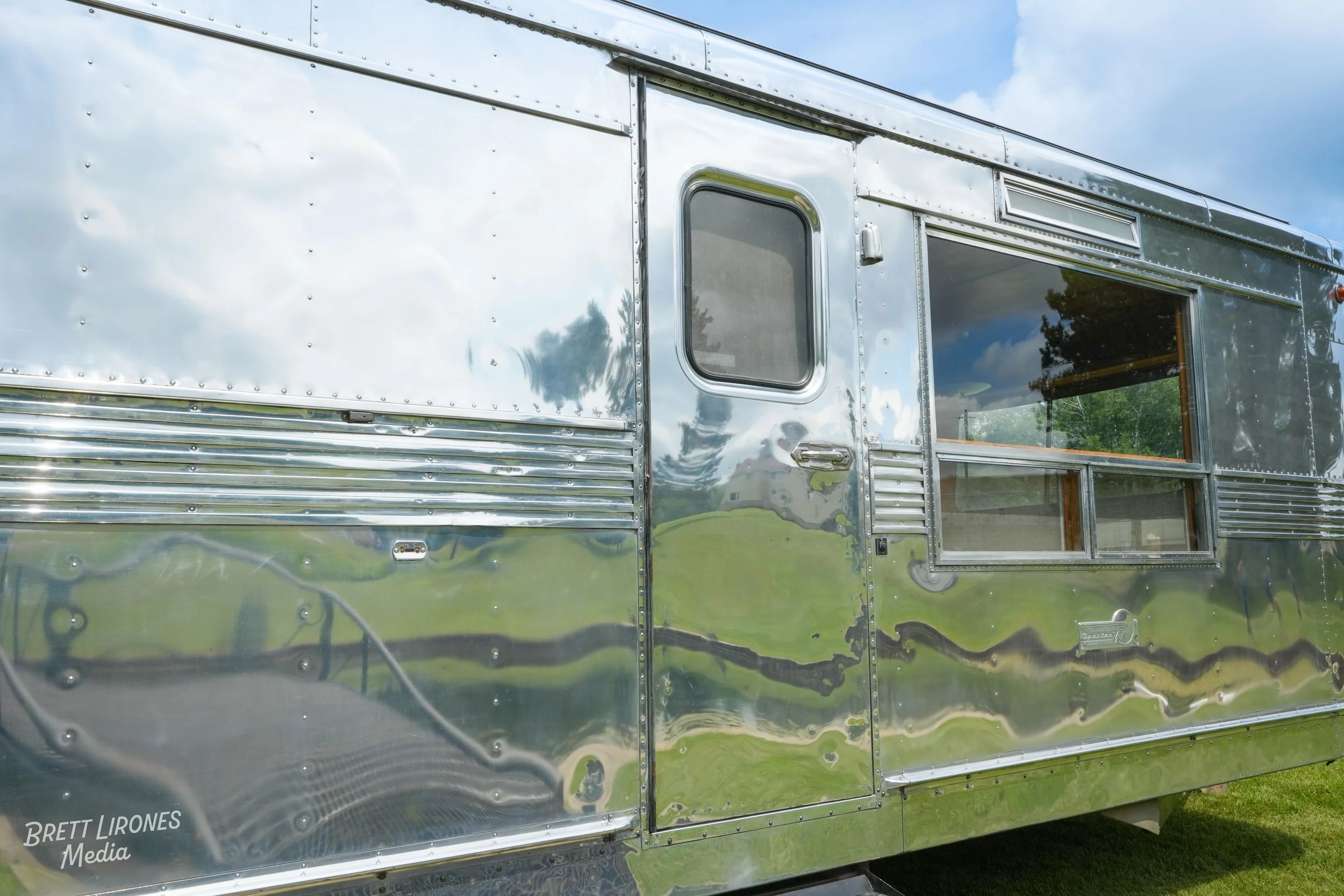 Close-up of a shiny, metallic food truck with a window and door, reflecting the sky and surrounding trees.