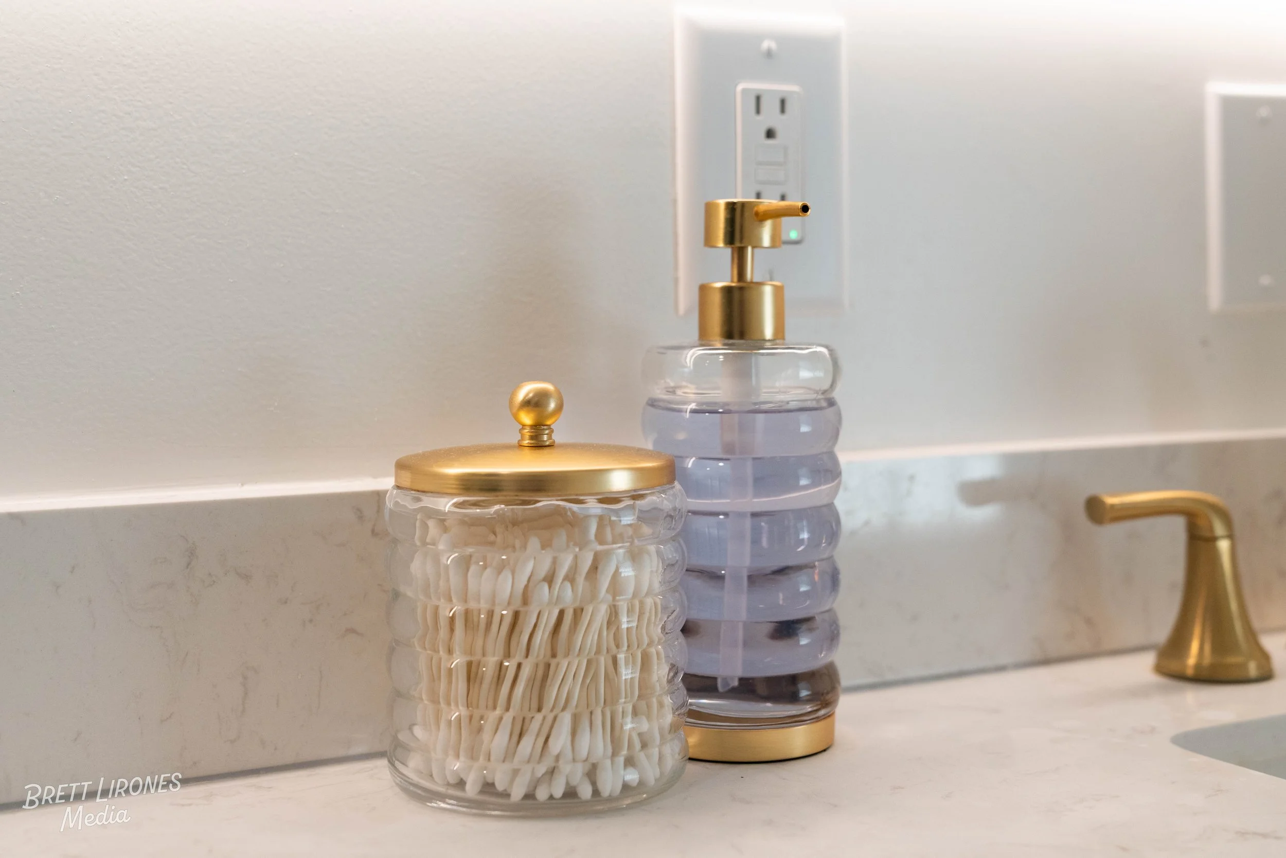 Bathroom countertop with two glass containers, one filled with cotton swabs and the other with liquid soap or lotion, all with gold-colored lids and pump, and a gold faucet on the sink.