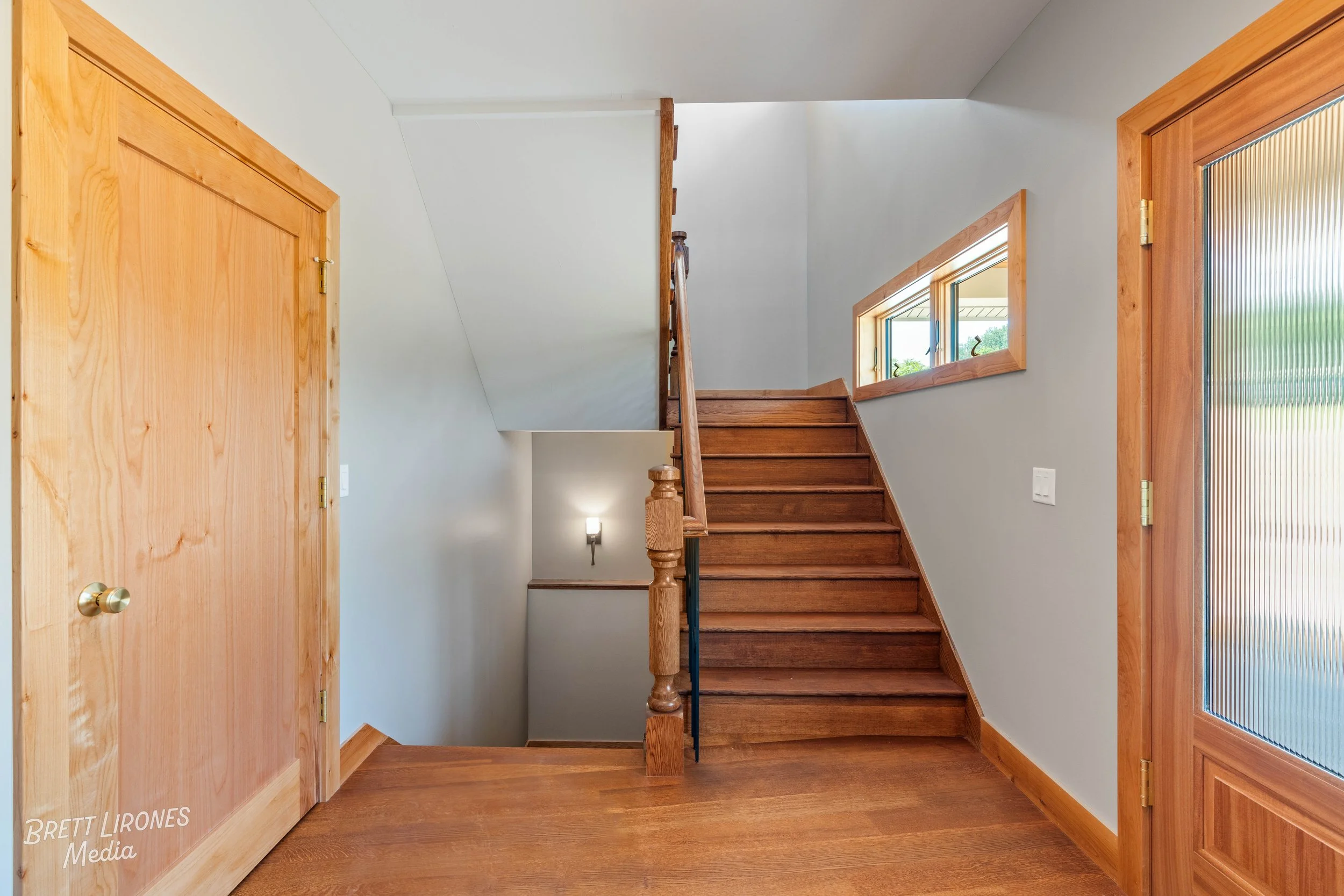 Interior view of a wooden staircase inside a house, with a small window on the right wall and a closed wooden door on the left. The staircase has a wooden railing and steps, with natural light coming through the window.