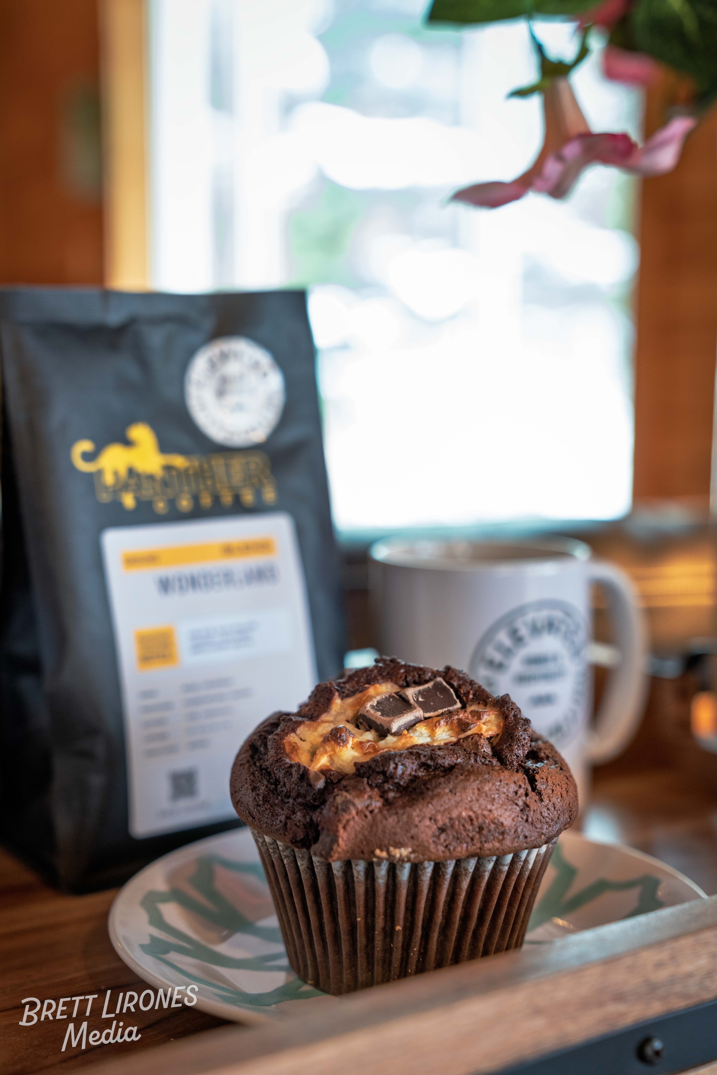 A chocolate muffin with marshmallows and chocolate pieces on top is on a decorative plate, with a coffee mug and a bag of coffee beans in the background.