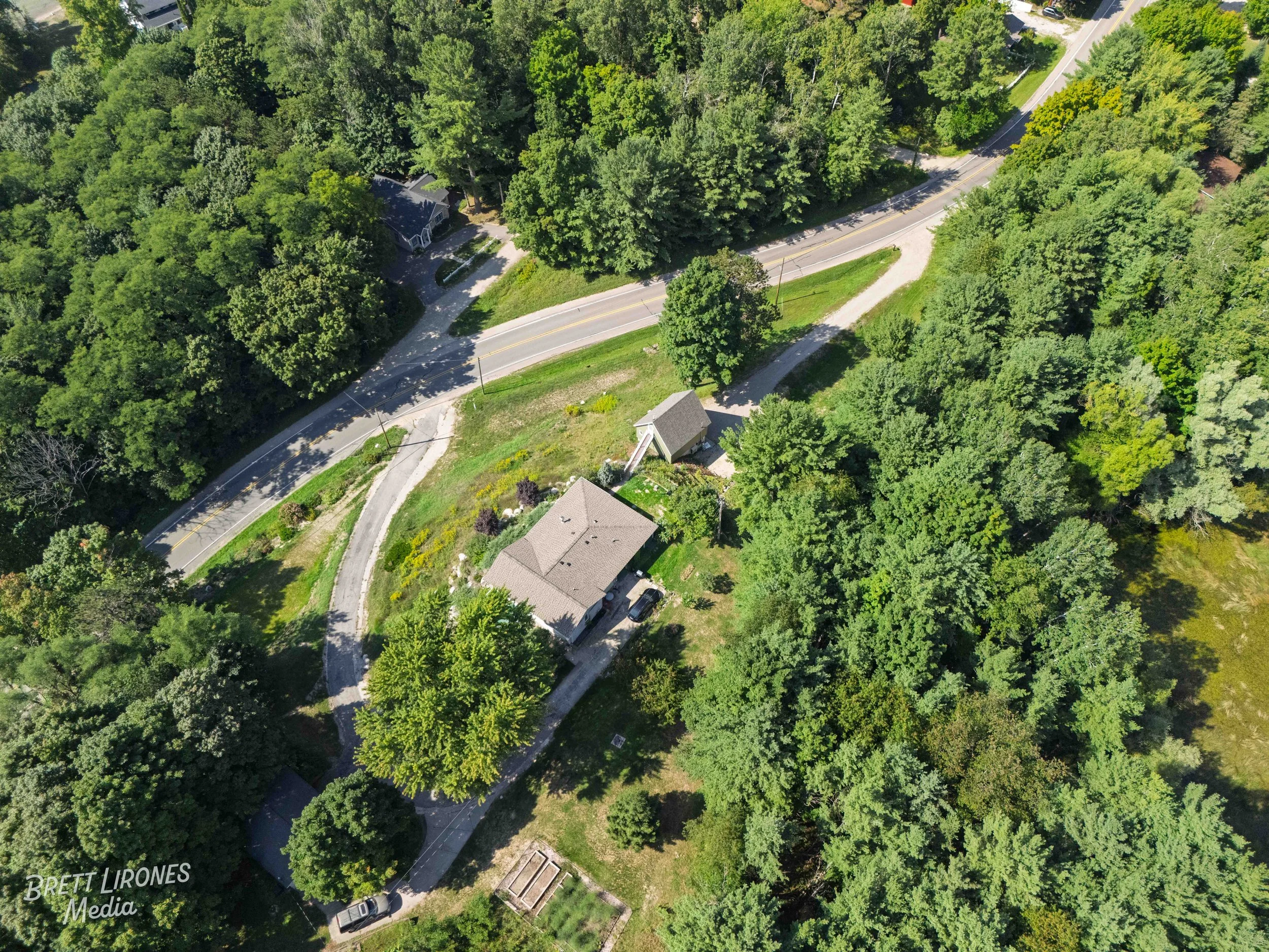 An aerial view of a house surrounded by trees, with a driveway leading to the house and a curved road nearby.