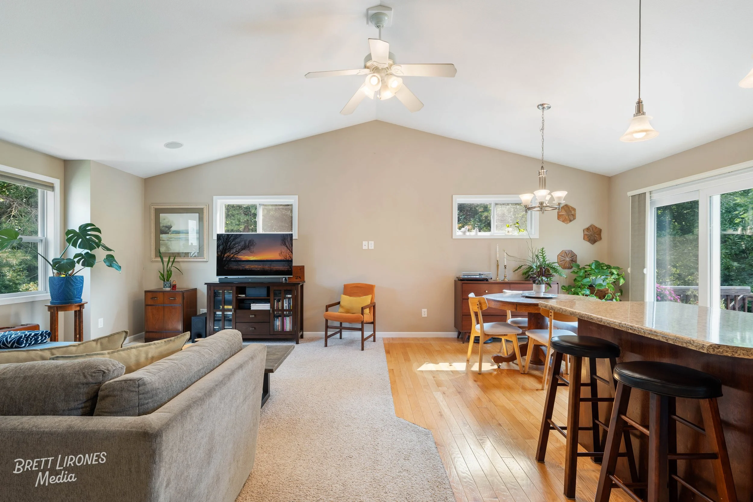 Living room with beige carpet and hardwood floor, beige walls, white ceiling with ceiling fan, large windows, TV on a wooden stand, sofa, chairs, and plants near sliding glass door.
