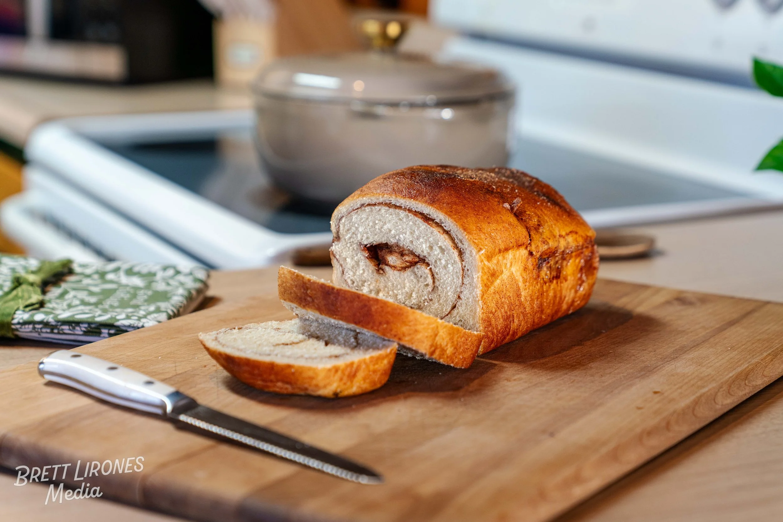 Half a cinnamon roll on a wooden cutting board with a serrated knife, in a kitchen setting