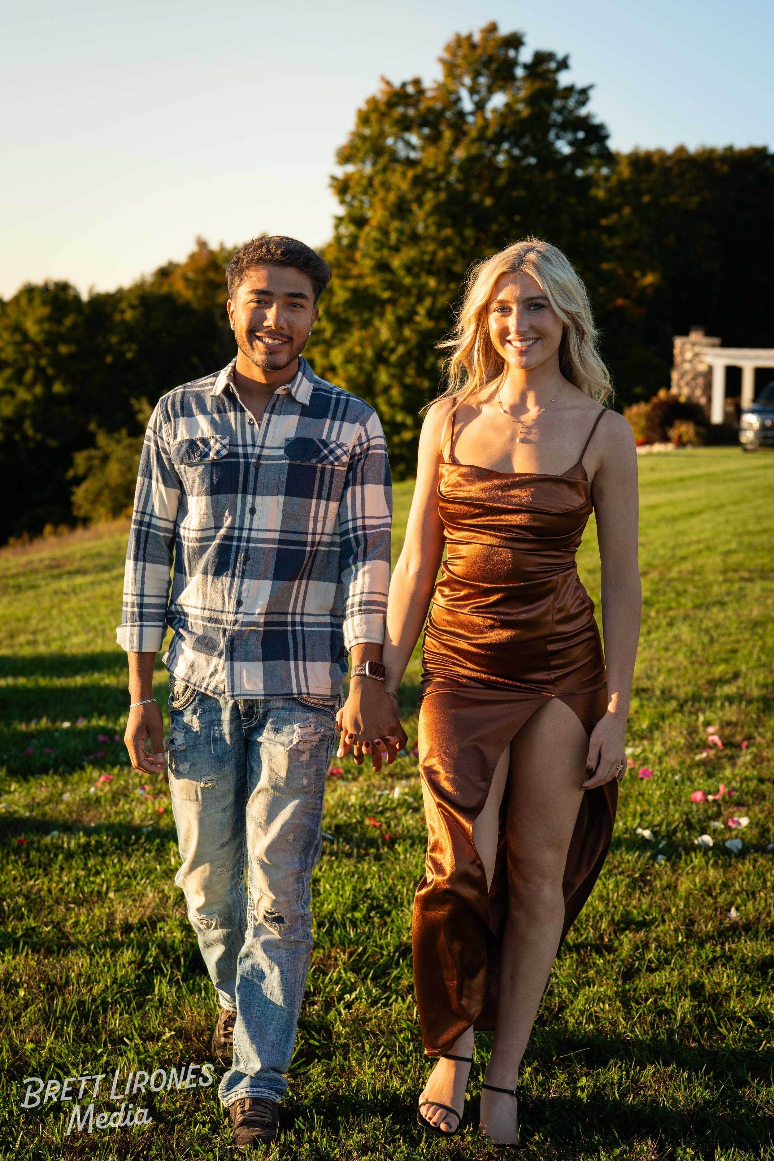 A couple holding hands walking on grass in a park during sunset. The man is wearing a plaid shirt and jeans, while the woman is dressed in a shiny, brown satin dress with a high slit and sandals. There are trees and a house in the background.