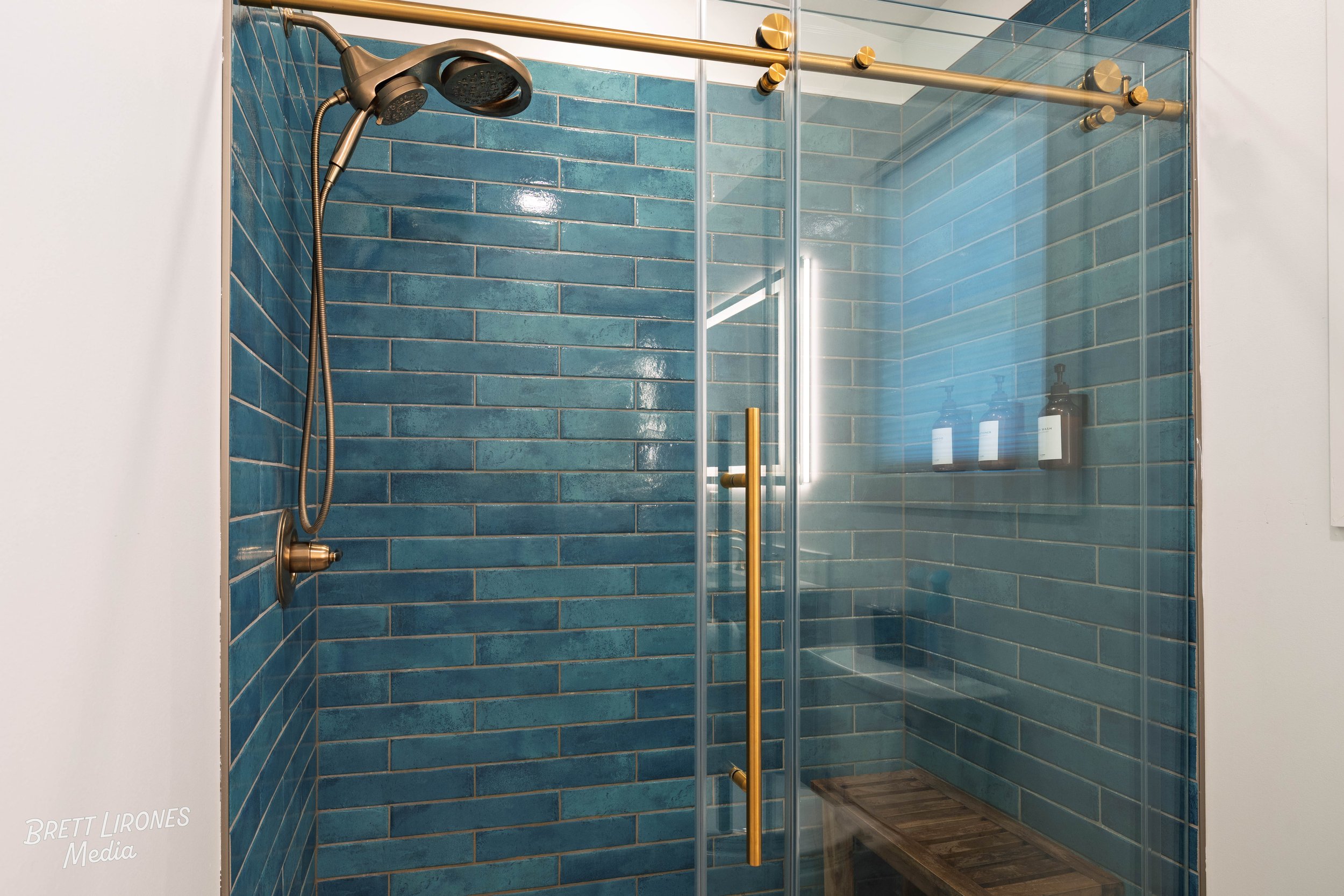 Modern glass-enclosed shower with blue subway tiles, featuring a gold handle and showerhead, with built-in niche containing bottles, and a wooden bench inside