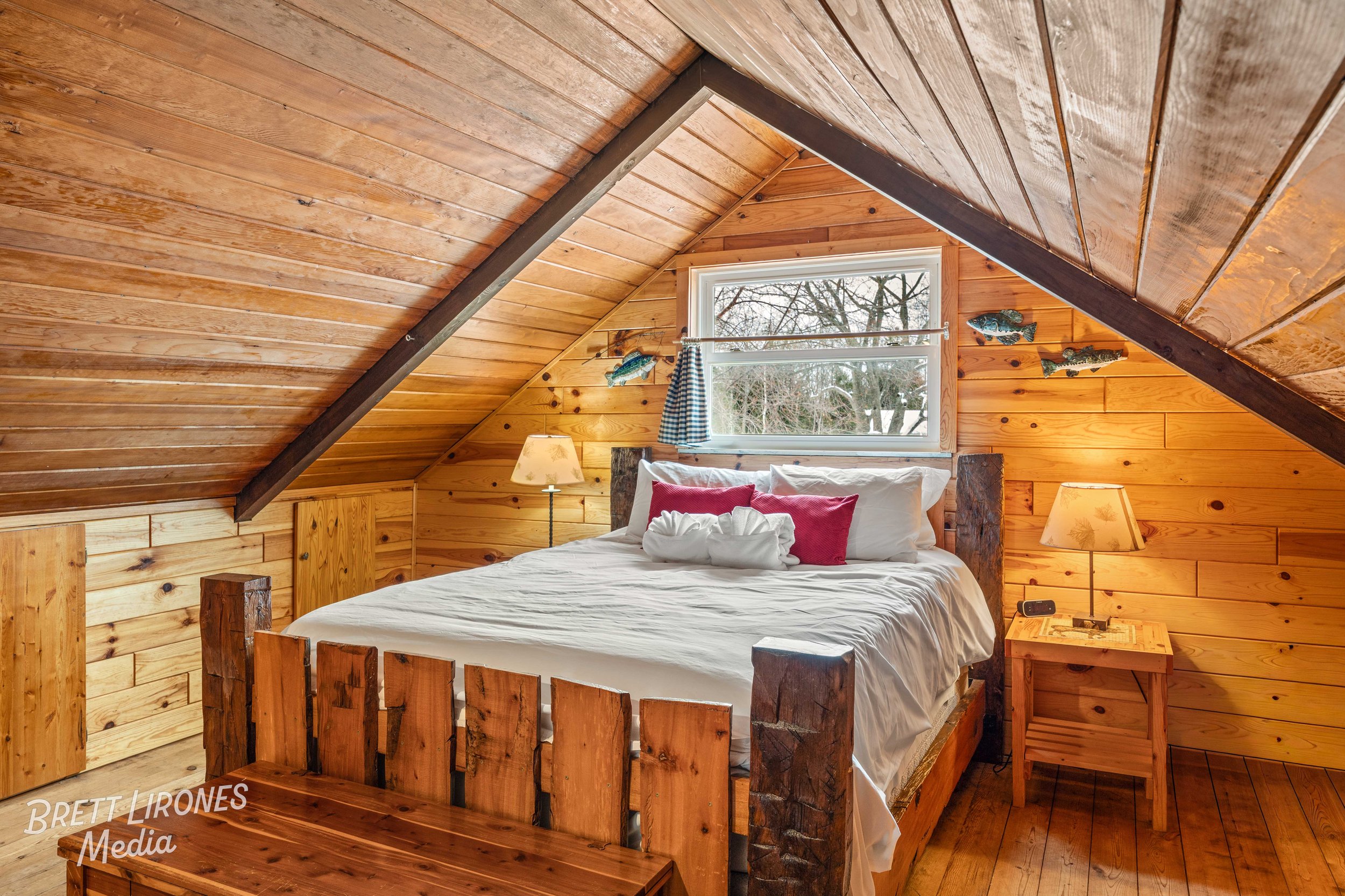 Cozy bedroom with wooden walls and sloped ceiling, featuring a bed with white linens and red pillows, two bedside lamps, and a window showing trees outside.