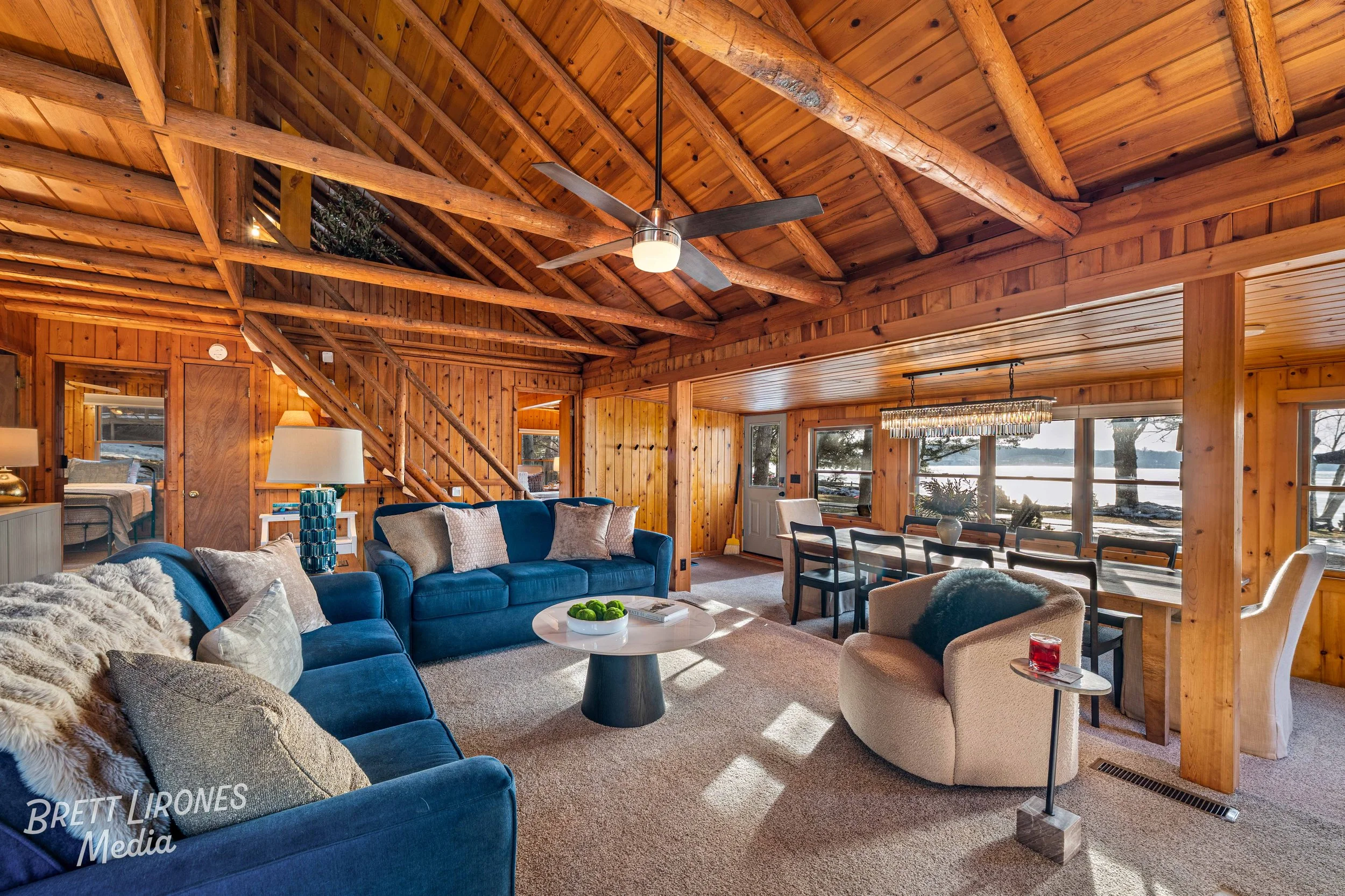Interior of a cozy wooden living room with large windows overlooking a lake, featuring blue and beige sofas, a round coffee table, and a dining area with a chandelier.
