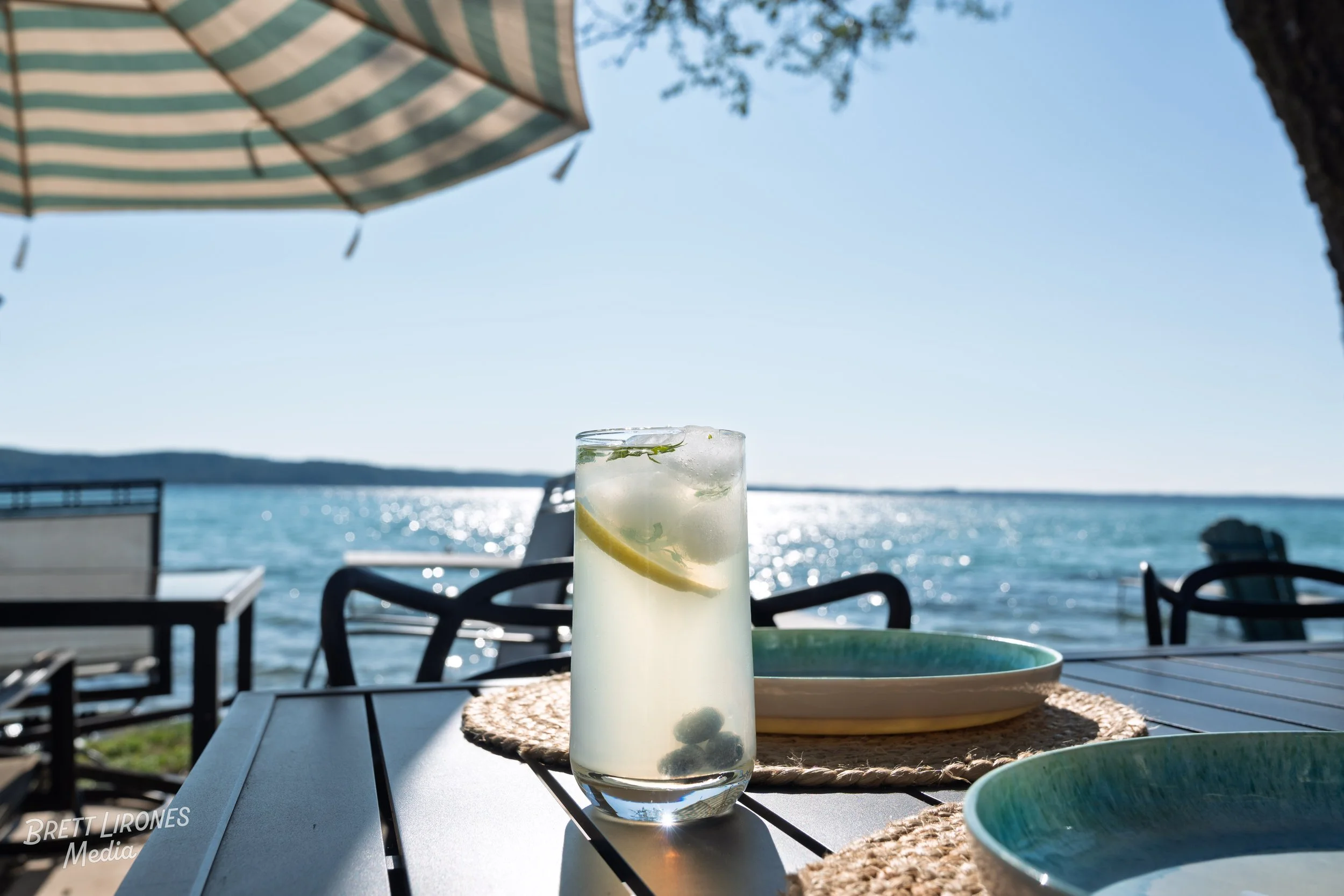 A glass of lemonade with lemon slices and mint leaves on a table by the water, with umbrellas, chairs, and a clear blue sky in the background.