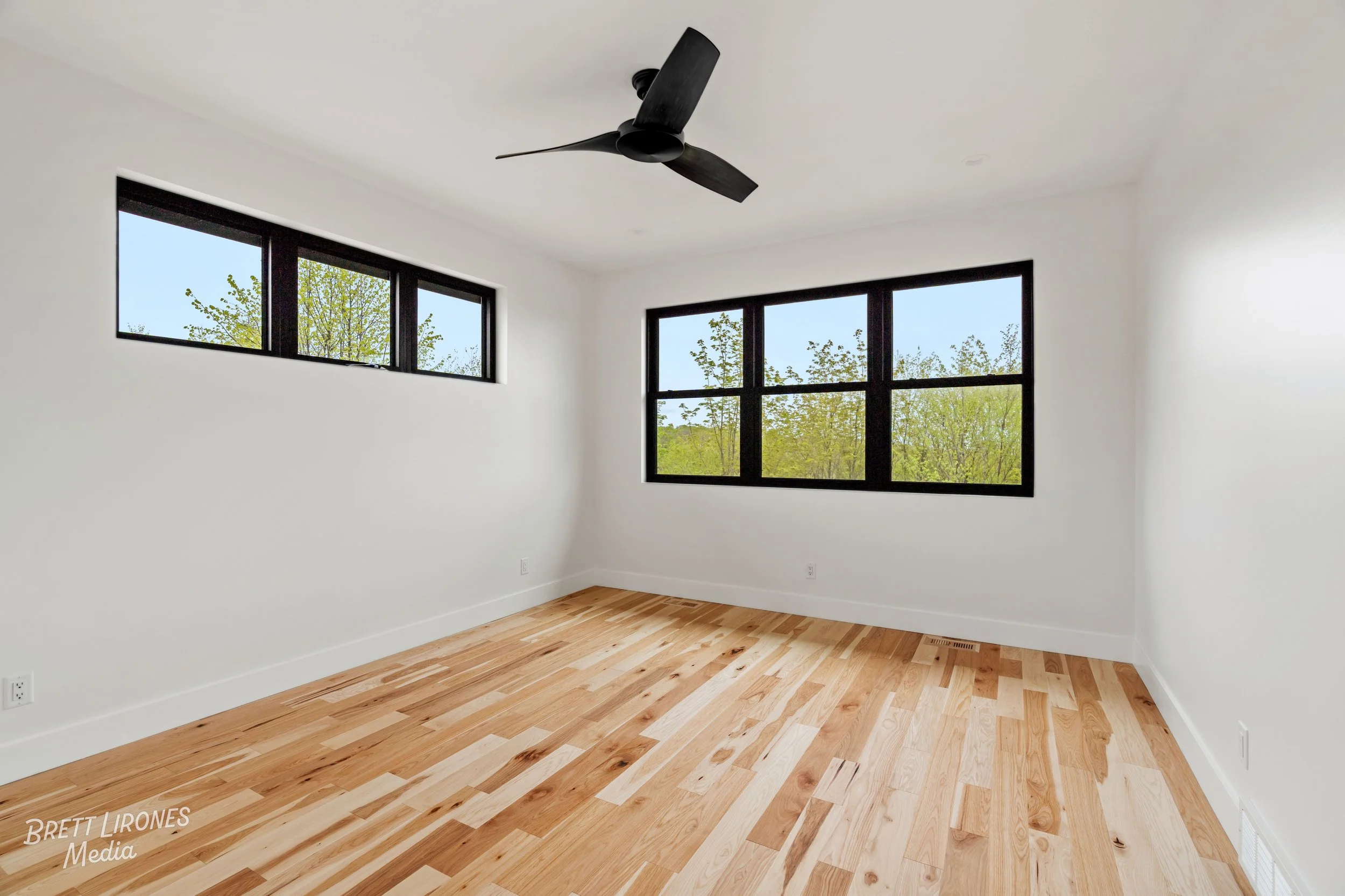Empty room with white walls, wooden floor, black-framed windows showing green trees outside, and ceiling fan.