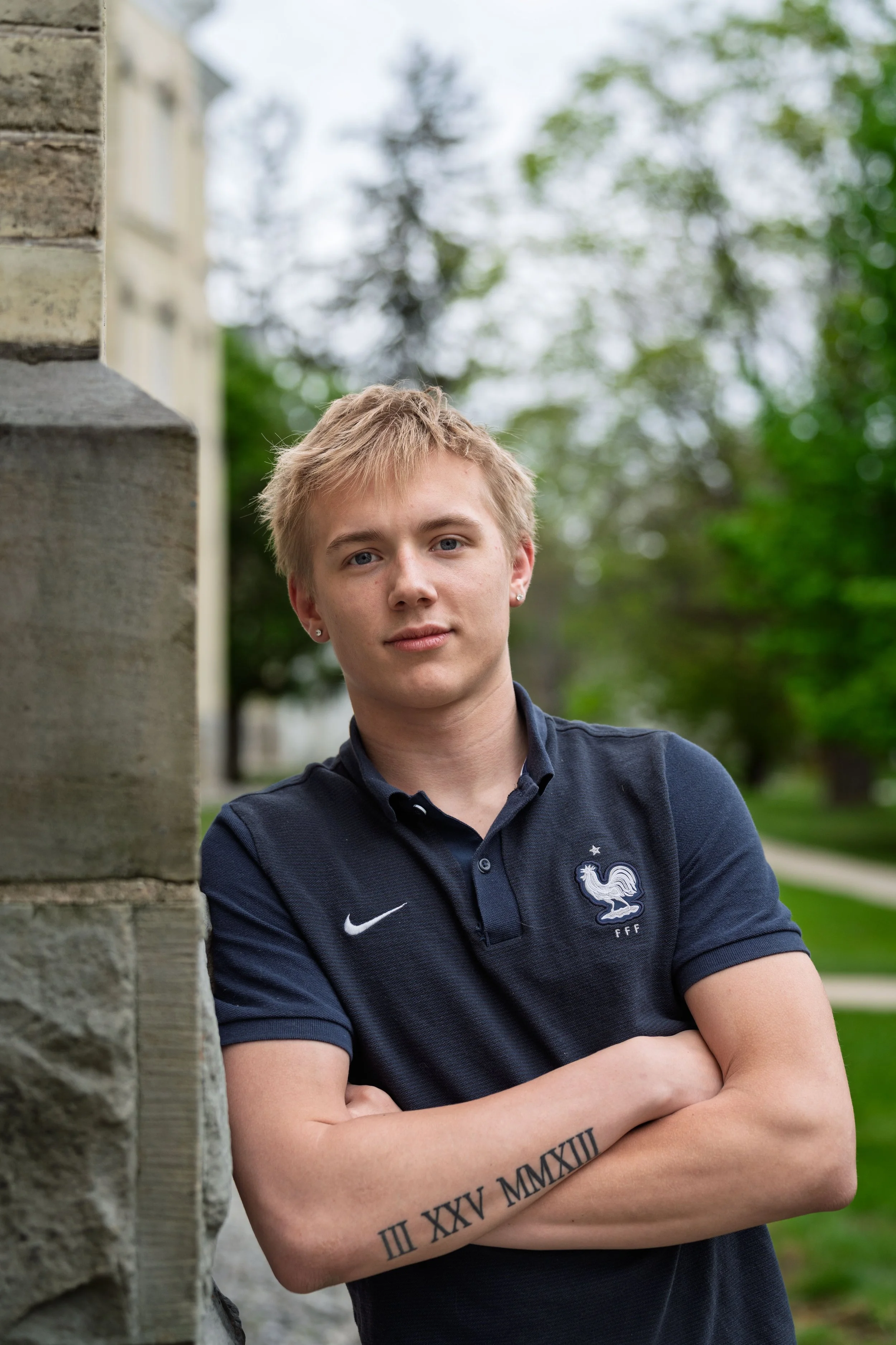 A young man with blond hair, dressed in a navy blue polo shirt with a French football logo, stands outdoors near a stone structure with crossed arms and a tattoo of Roman numerals on his forearm, with a background of trees and greenery.